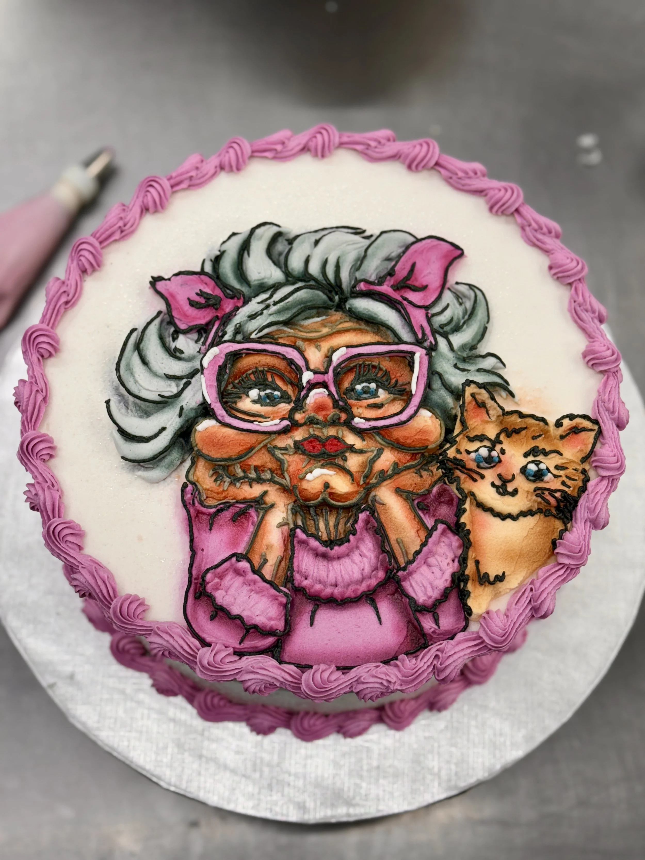 Decorative cake with a detailed icing illustration of a woman with glasses, gray hair with pink accents, and a kitten, all with expressive eyes and intricate details, surrounded by a pink piped border.