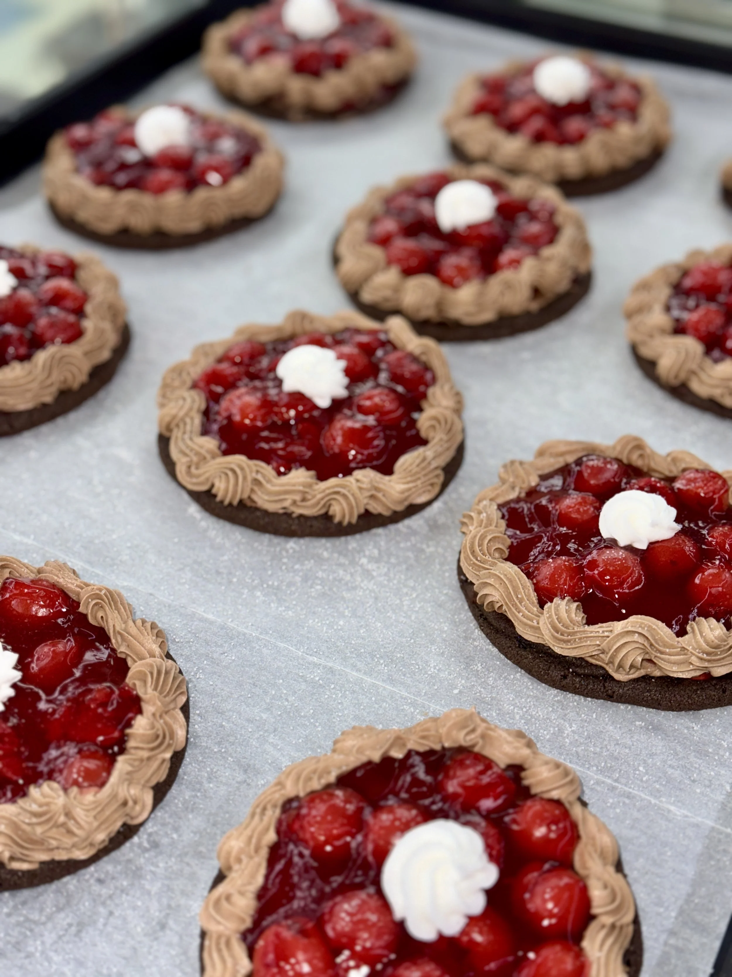 Chocolate cookies topped with red fruit jam and a dollop of white cream, decorated with chocolate piped borders.