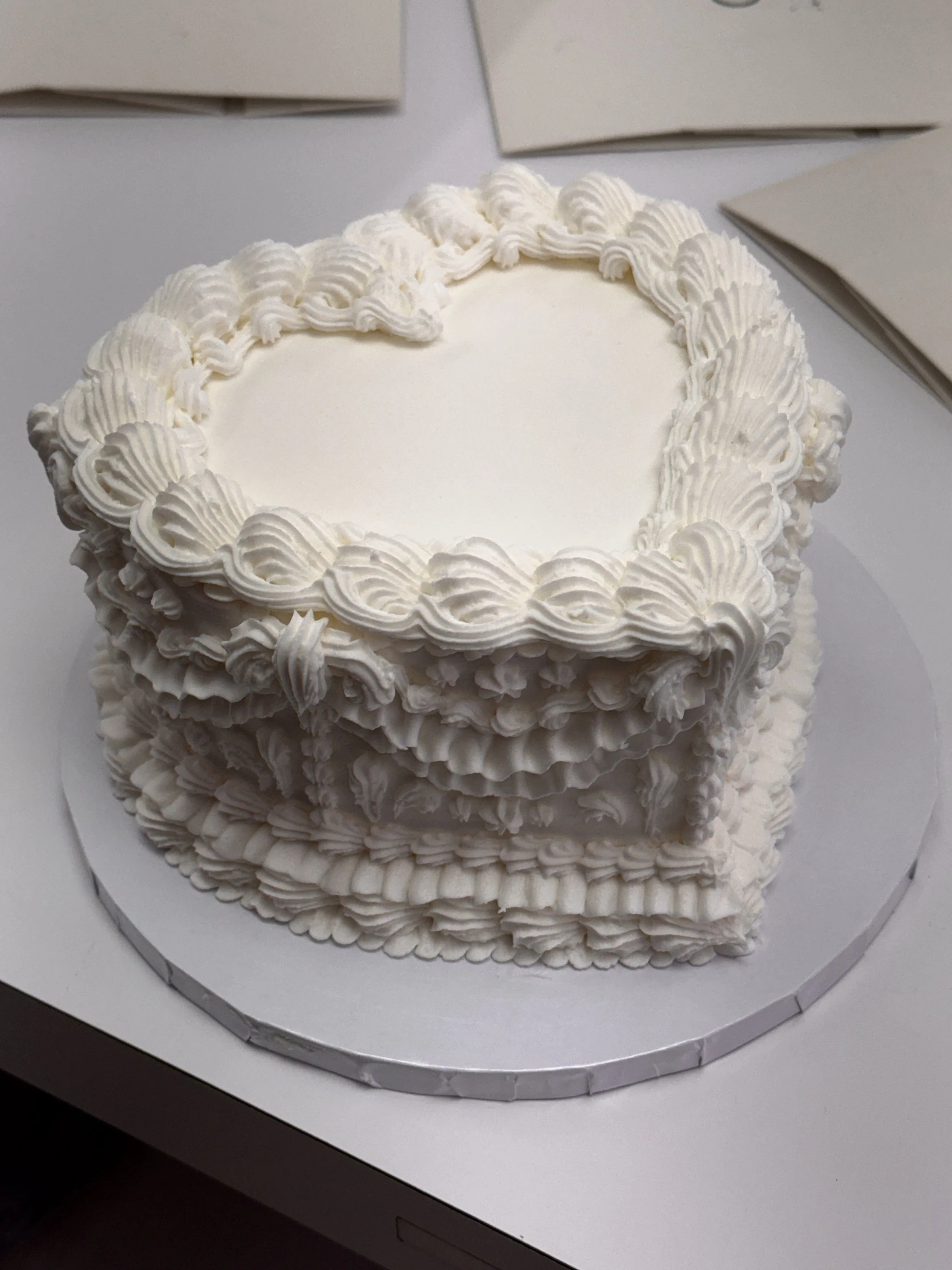 A round white cake decorated with white frosting in a shell design, sitting on a white surface with cardboard boxes in the background.