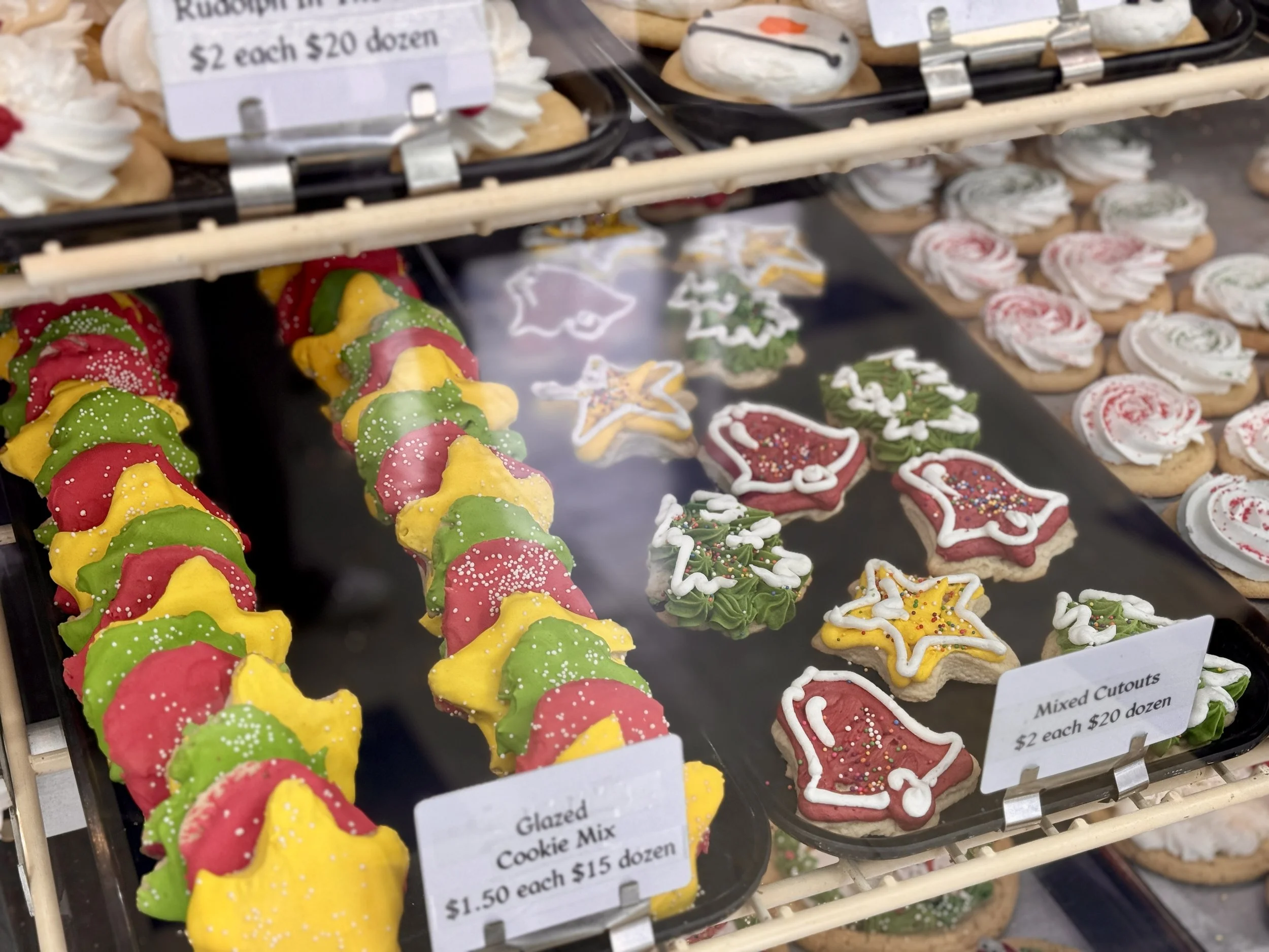 Display of decorated Christmas cookies on a bakery tray, including Christmas trees, stockings, and star shapes, decorated with icing and colorful sprinkles.