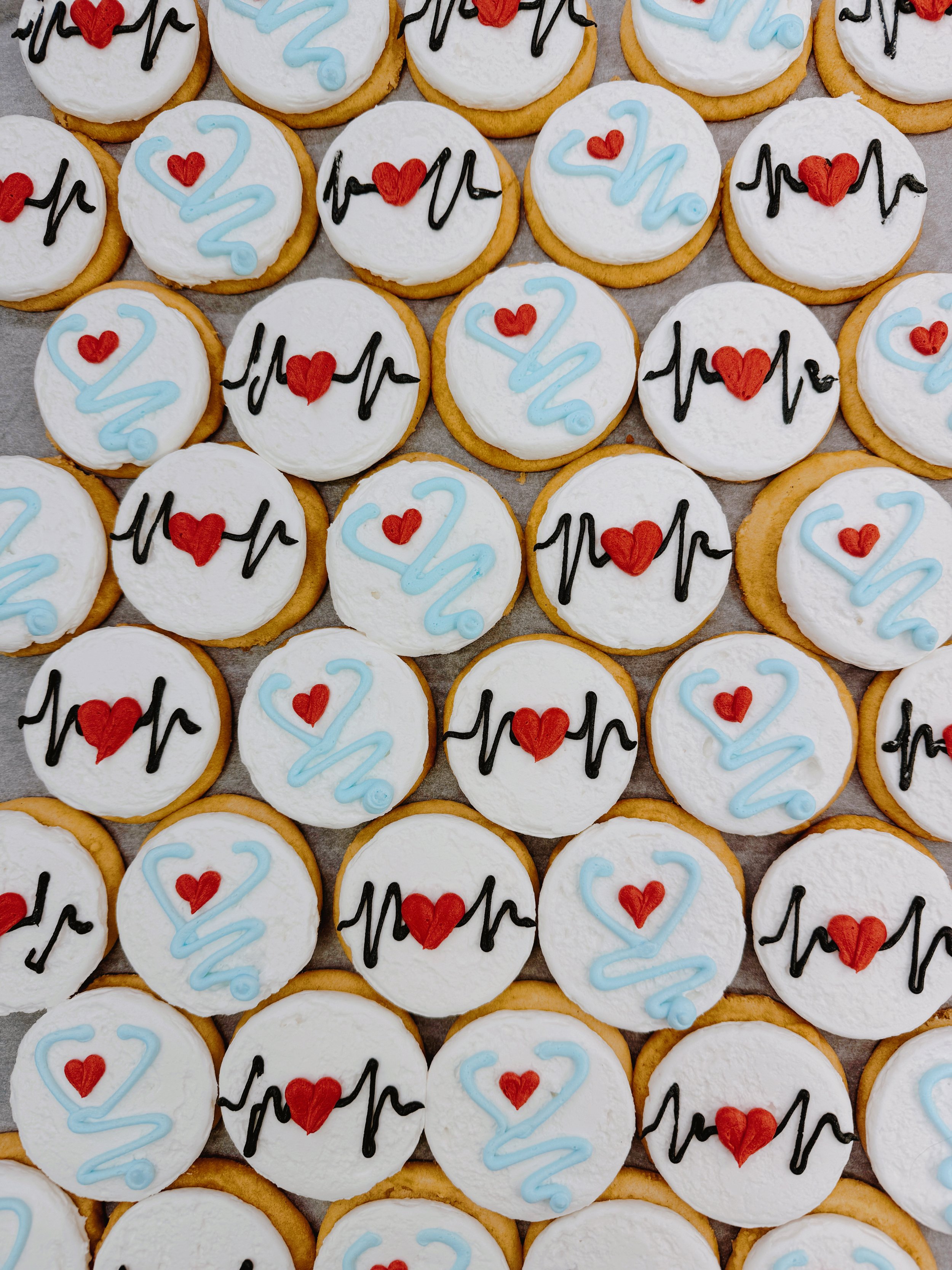 Assorted round cookies decorated with white icing, red hearts, and black and blue squiggly lines, arranged in a grid.