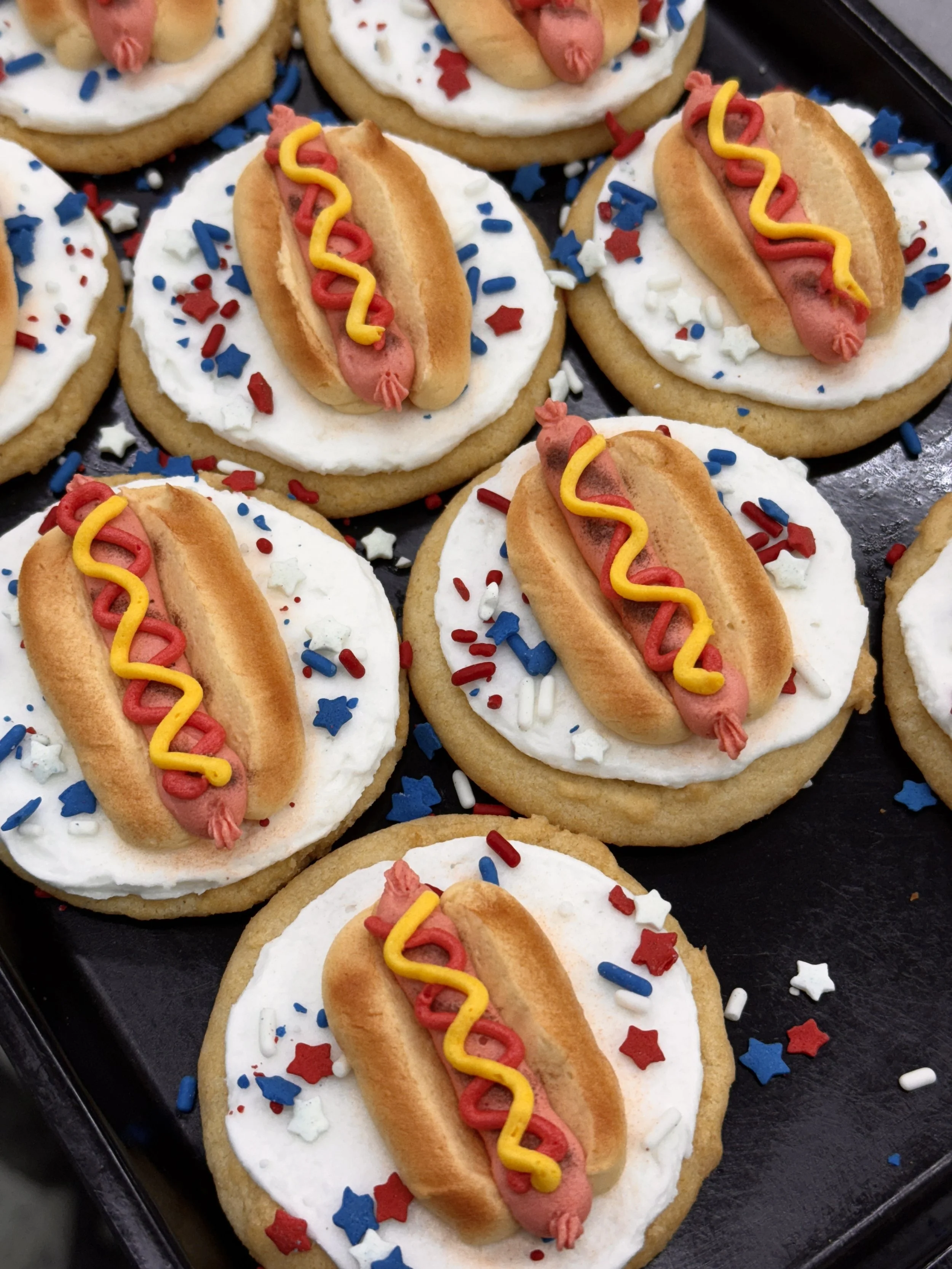 Hot dog cookies decorated with white icing, red, yellow, and blue sprinkles, resembling hot dogs in buns.