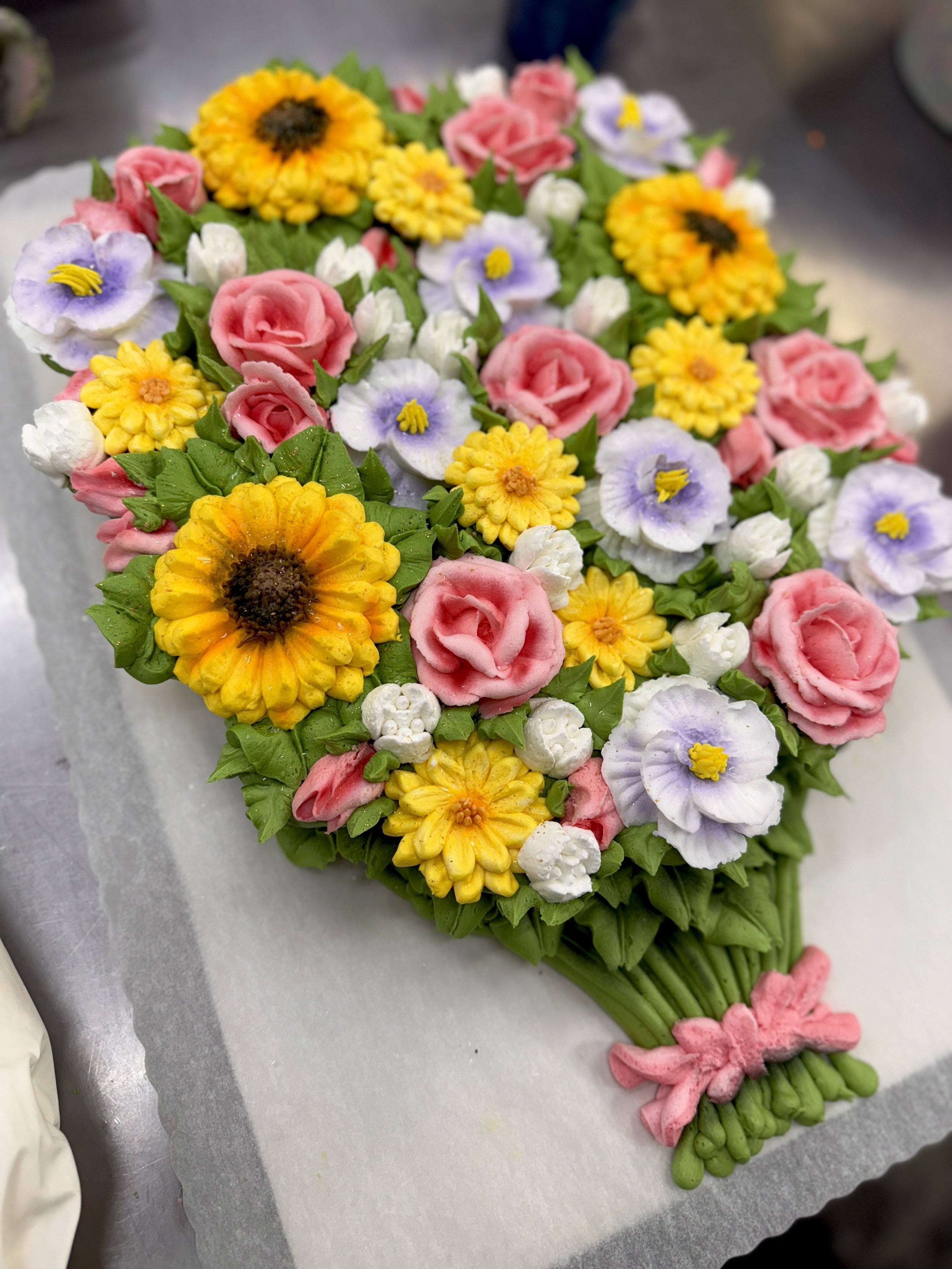 Colorful floral arrangement with pink roses, yellow marigolds, purple and white pansies, and green foliage, shaped like an elongated bouquet.