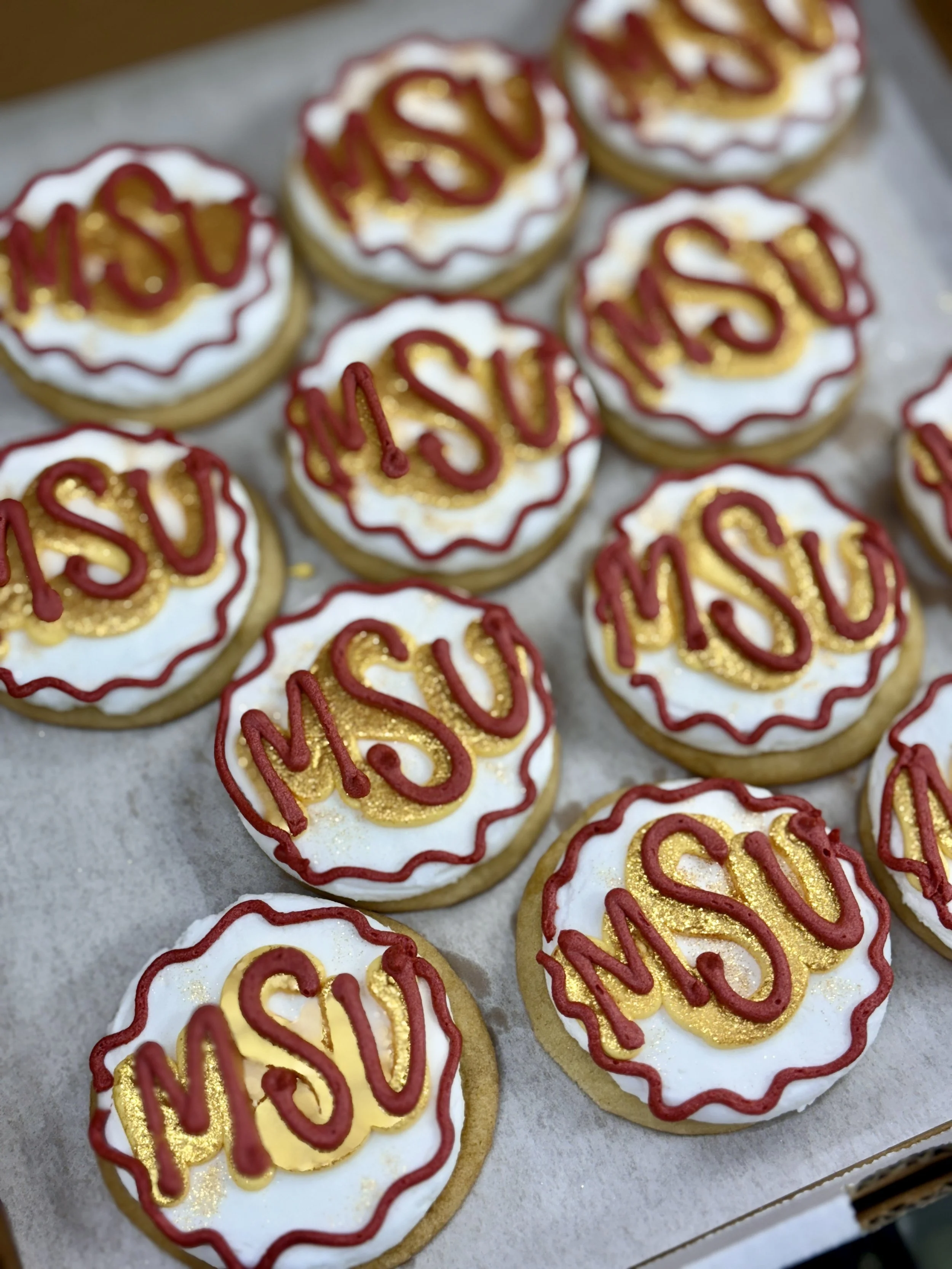 Decorated cookies with white icing, red and gold writing that says 'MOM' or 'MRS,' with red and gold outlining, on a baking sheet lined with parchment paper.