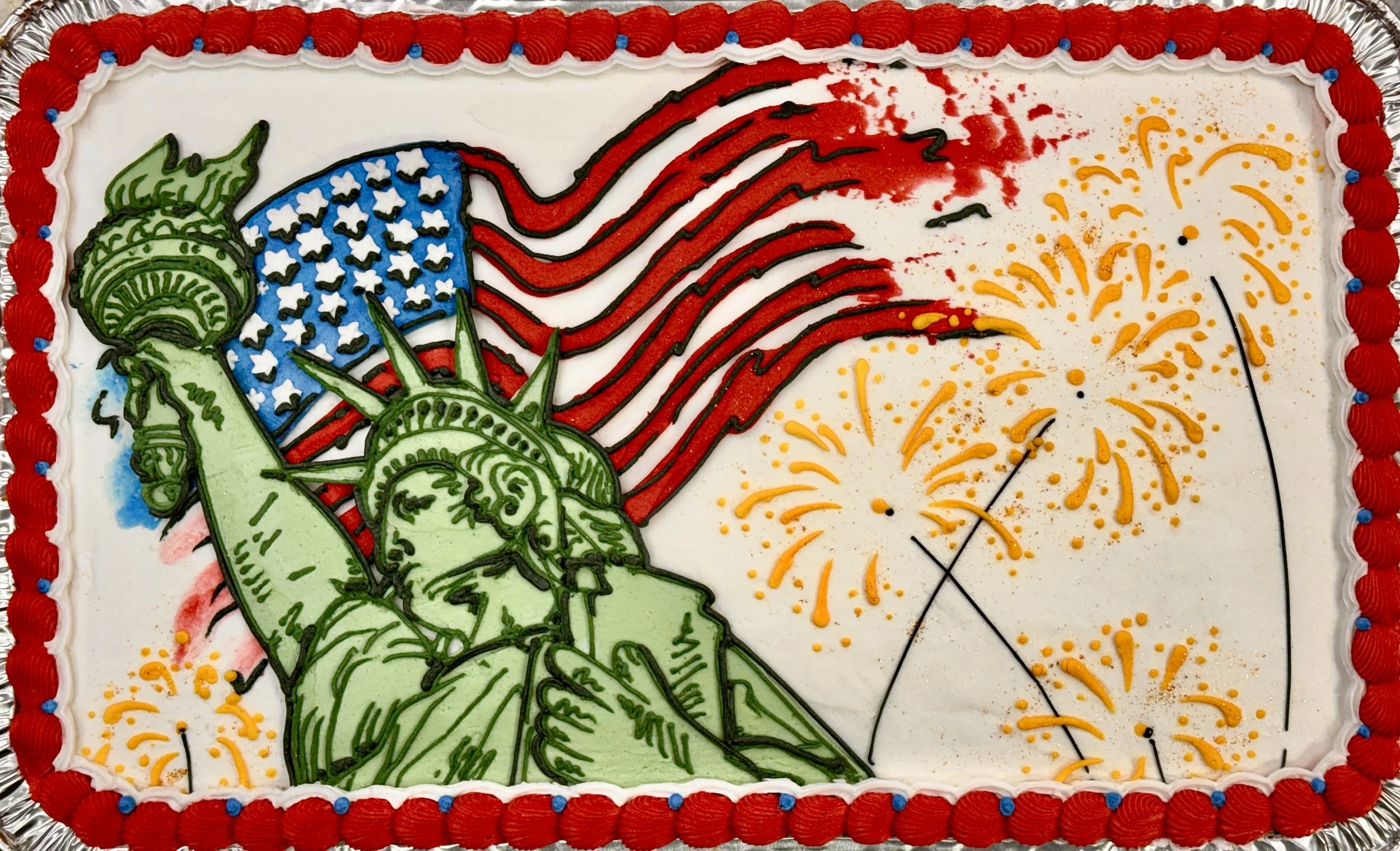 A decorated rectangular cake featuring the Statue of Liberty and an American flag with red, white, blue, yellow, and black icing details, surrounded by a red border with blue accents.
