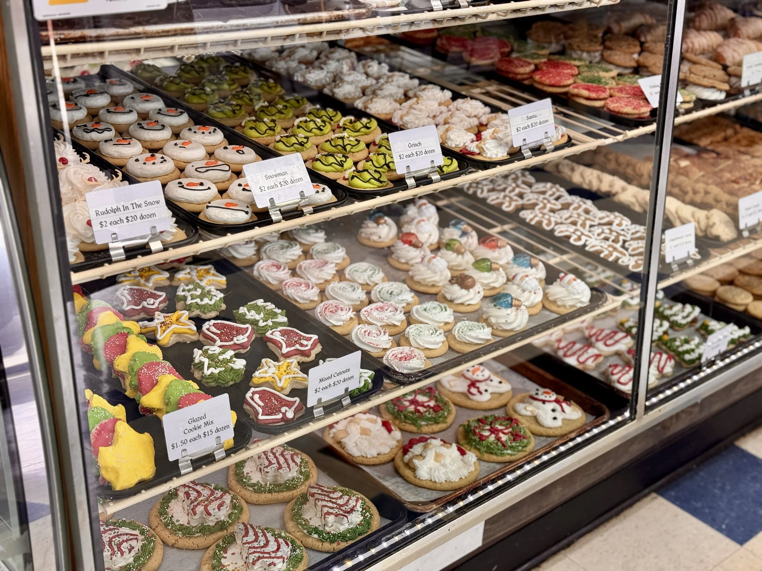A display case filled with decorated holiday cookies, including snowmen, Christmas trees, and other festive shapes and patterns, with price tags for individual cookies and dozen quantities.