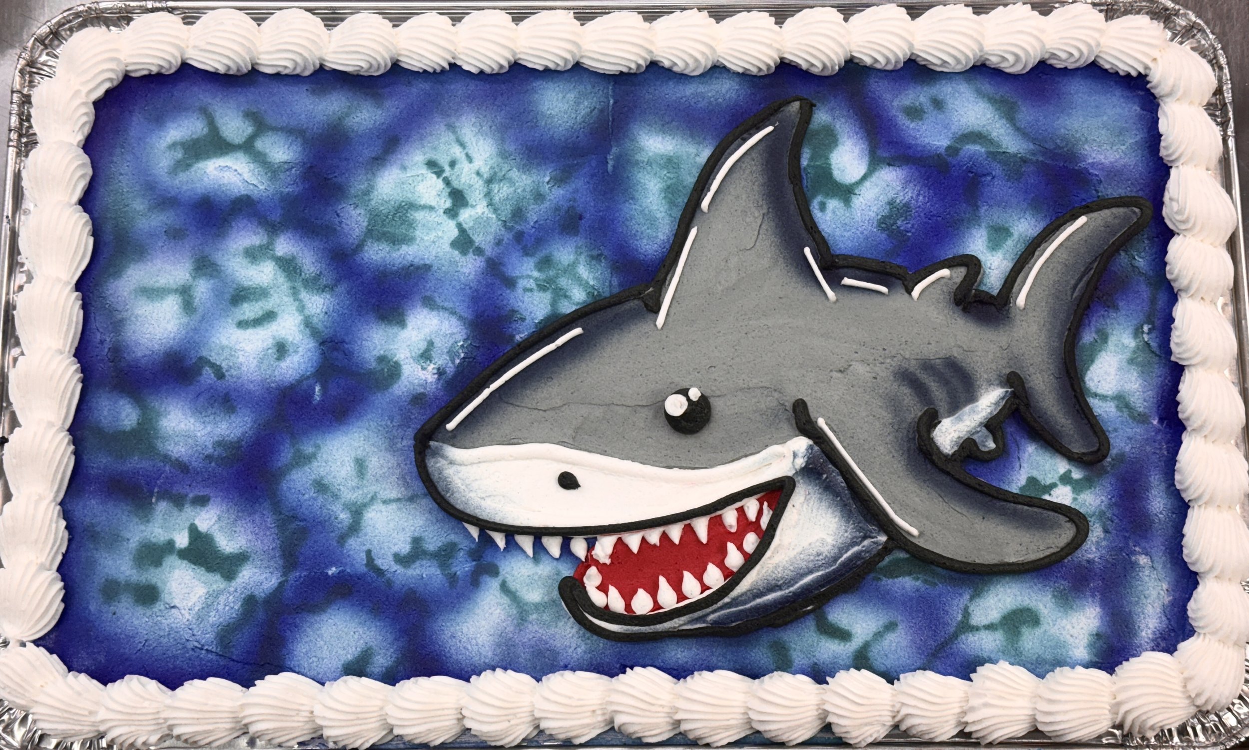 A decorated birthday cake featuring a shark motif with blue, green, and purple swirled frosting background and a cartoonish shark face with open mouth, sharp teeth, and expressive eyes, bordered with white frosting.