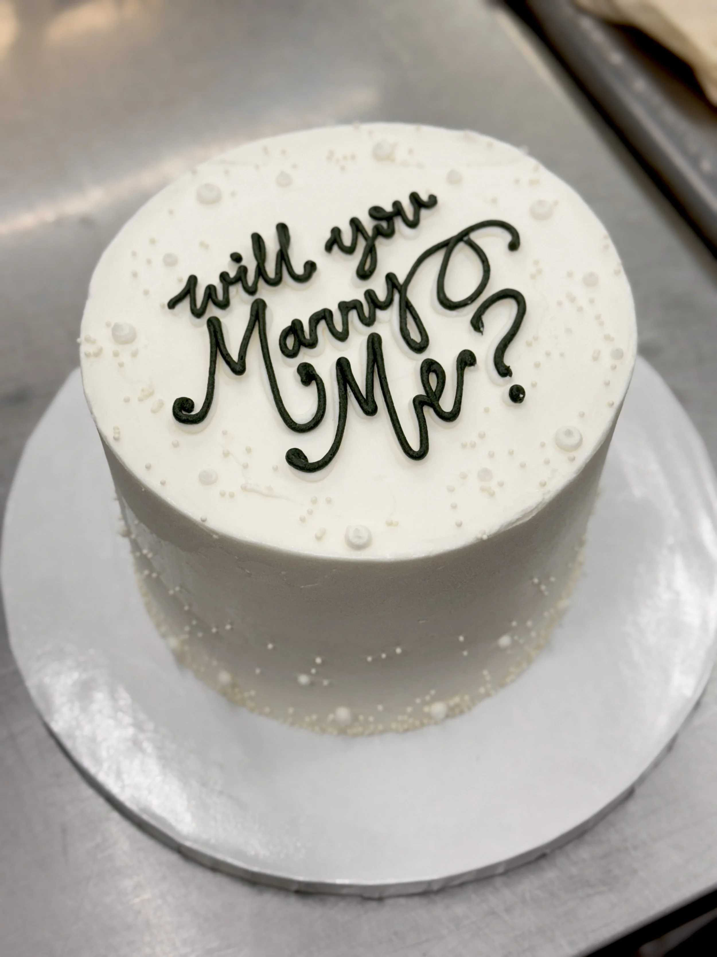 A round white cake with black icing writing that says "Will you marry me?"