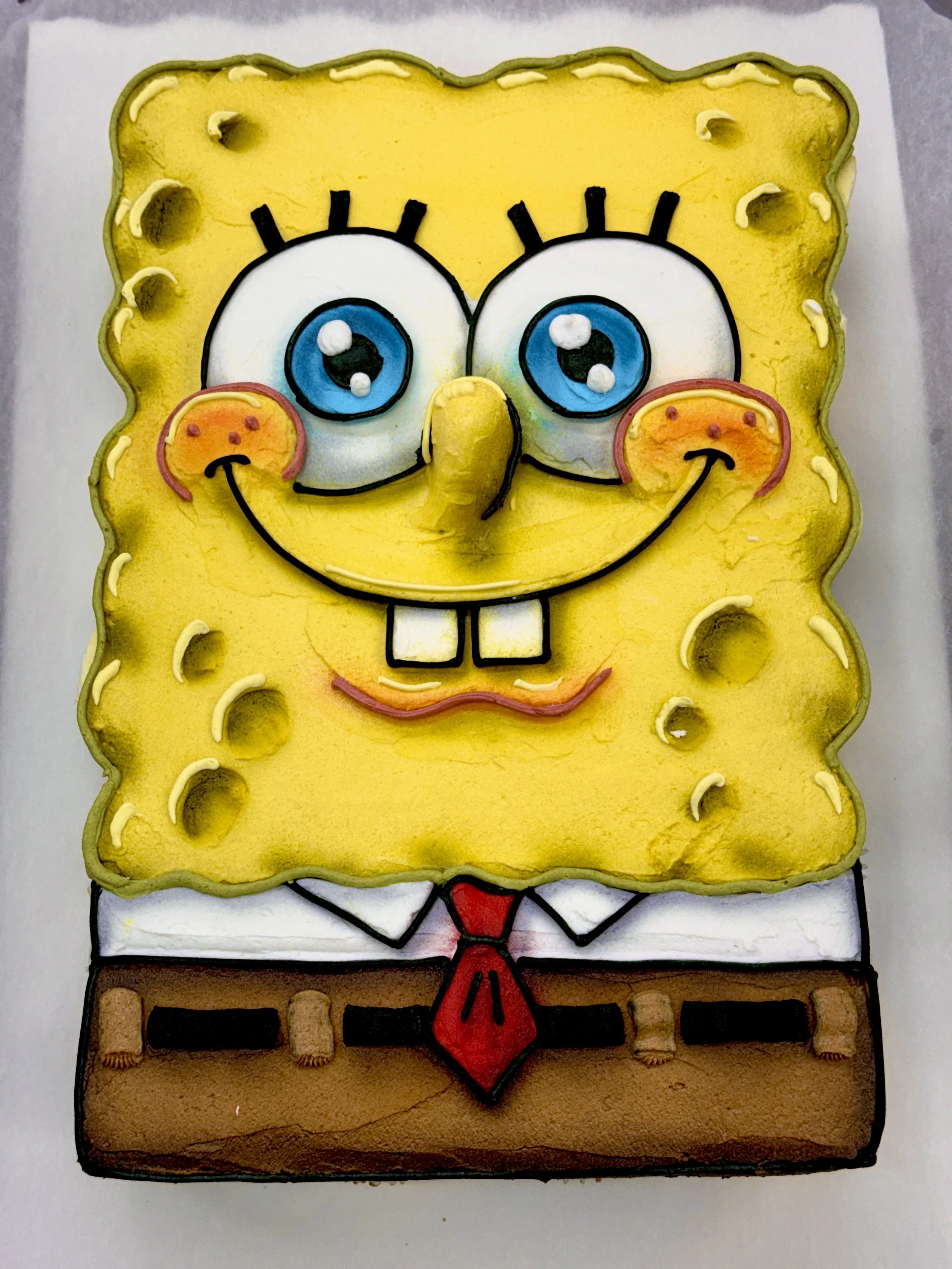 Cake in the shape of SpongeBob SquarePants with blue eyes, brown pants, and red tie, decorated with yellow icing, fondant, and various edible details.