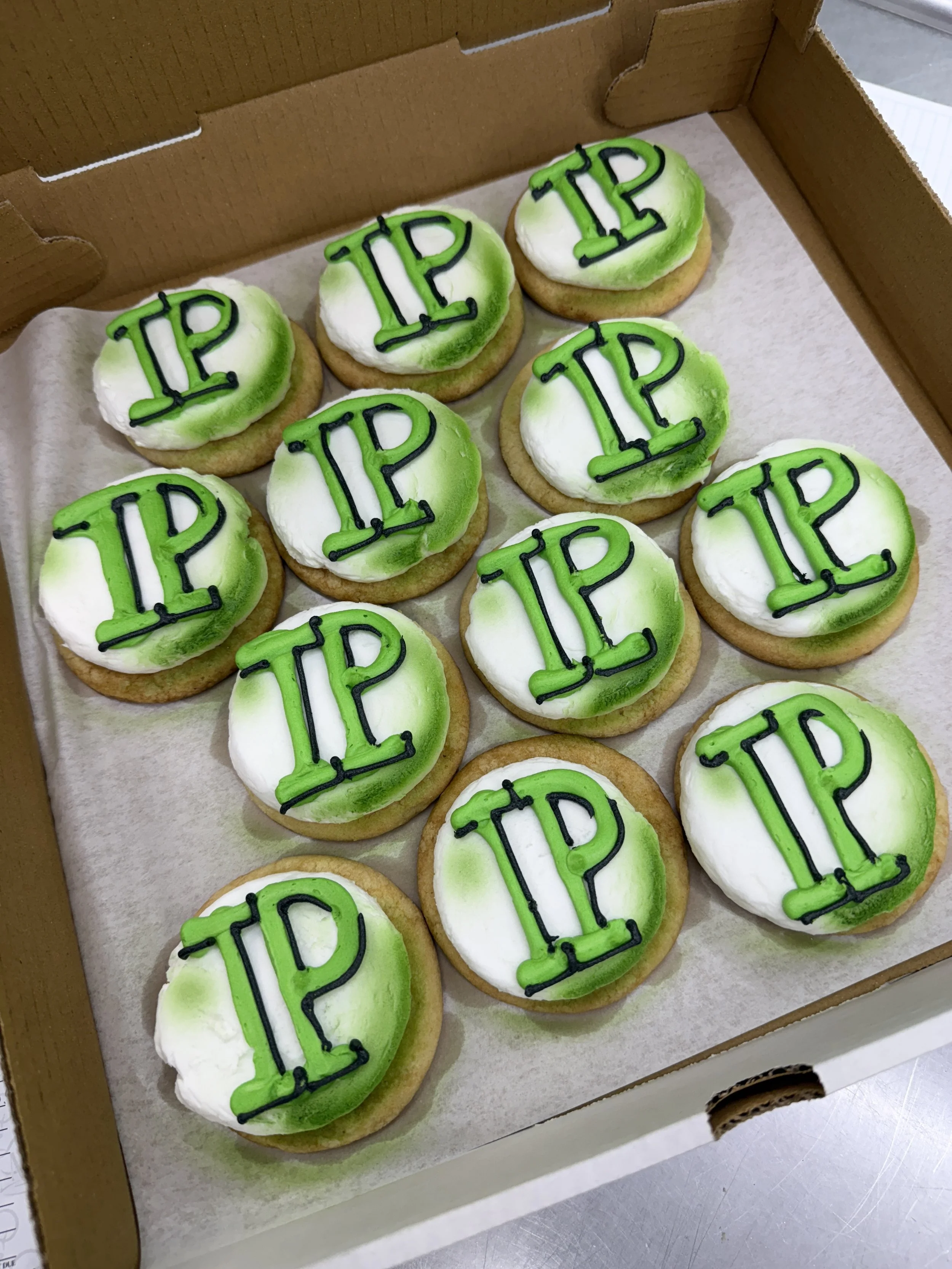 A box of decorated cookies with white and green icing, each featuring the word 'TIP' in black icing outlined with green, and arranged in two rows inside a cardboard box lined with parchment paper.