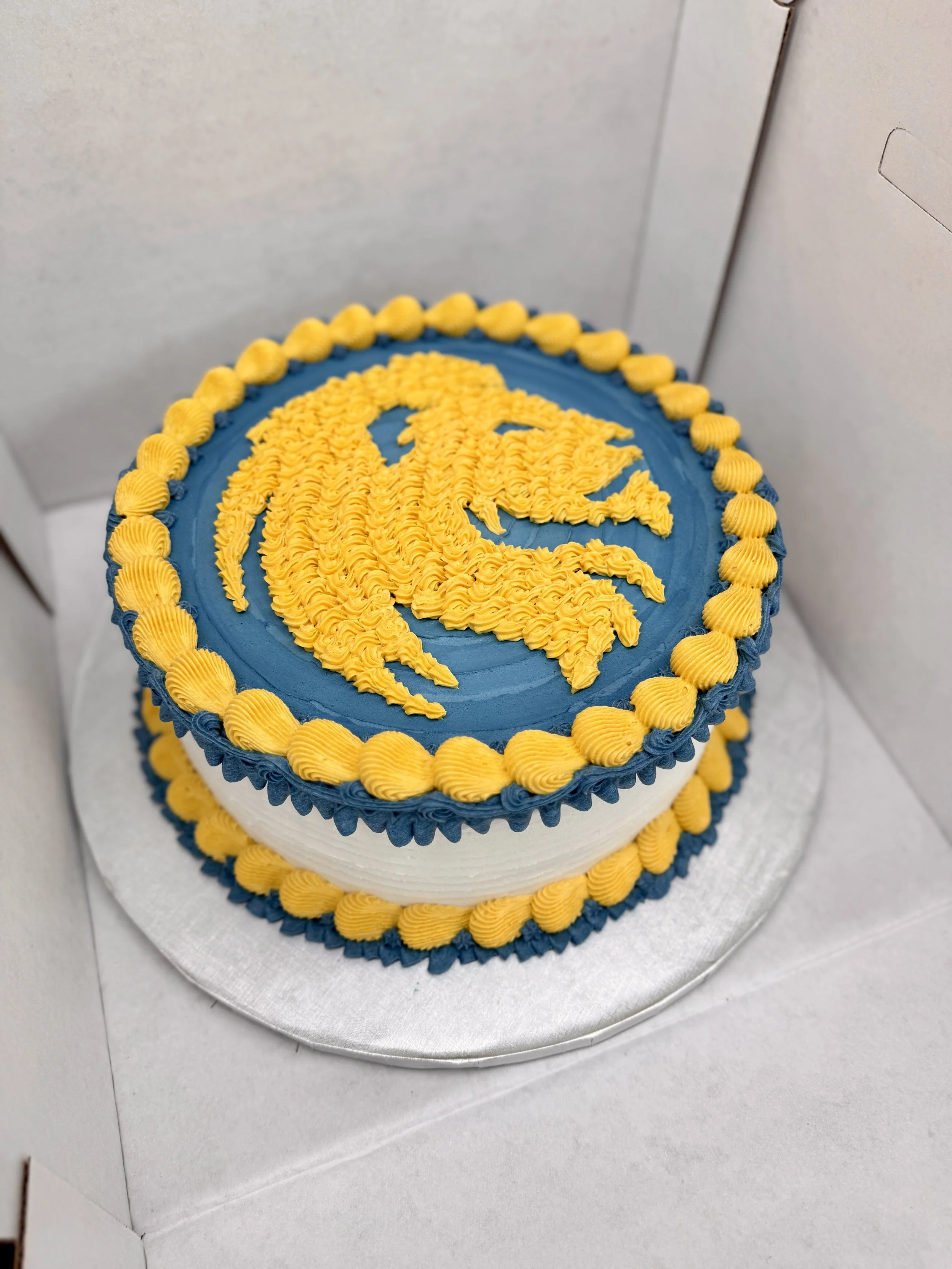 A round cake with blue and white frosting, decorated with a yellow crest and border made of yellow and blue piping, placed on a silver board inside a white box.