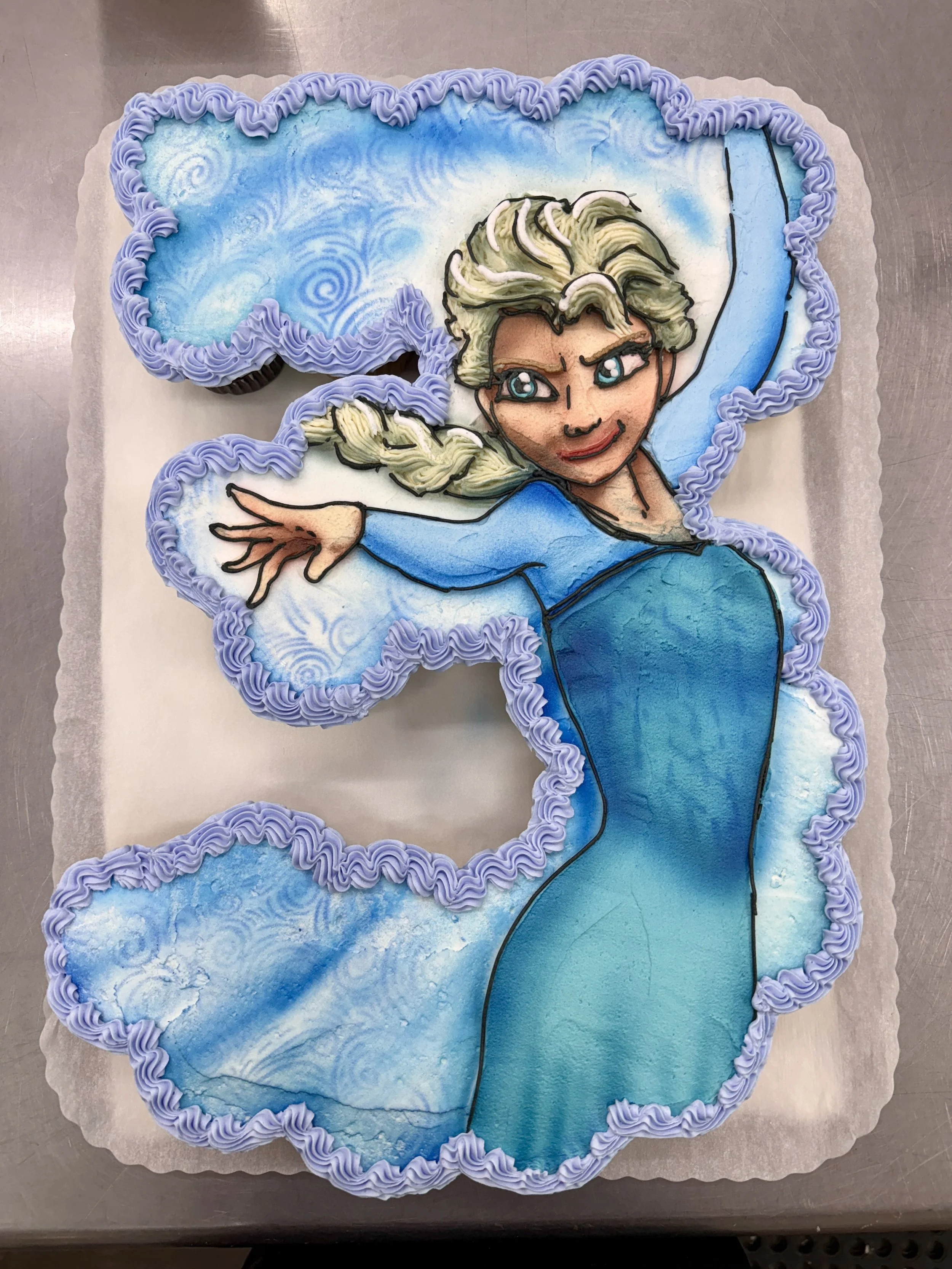A large decorative cake in the shape of Elsa from Frozen, featuring her face, hair, and ice powers, with light purple piped border and swirling blue and white icy background.