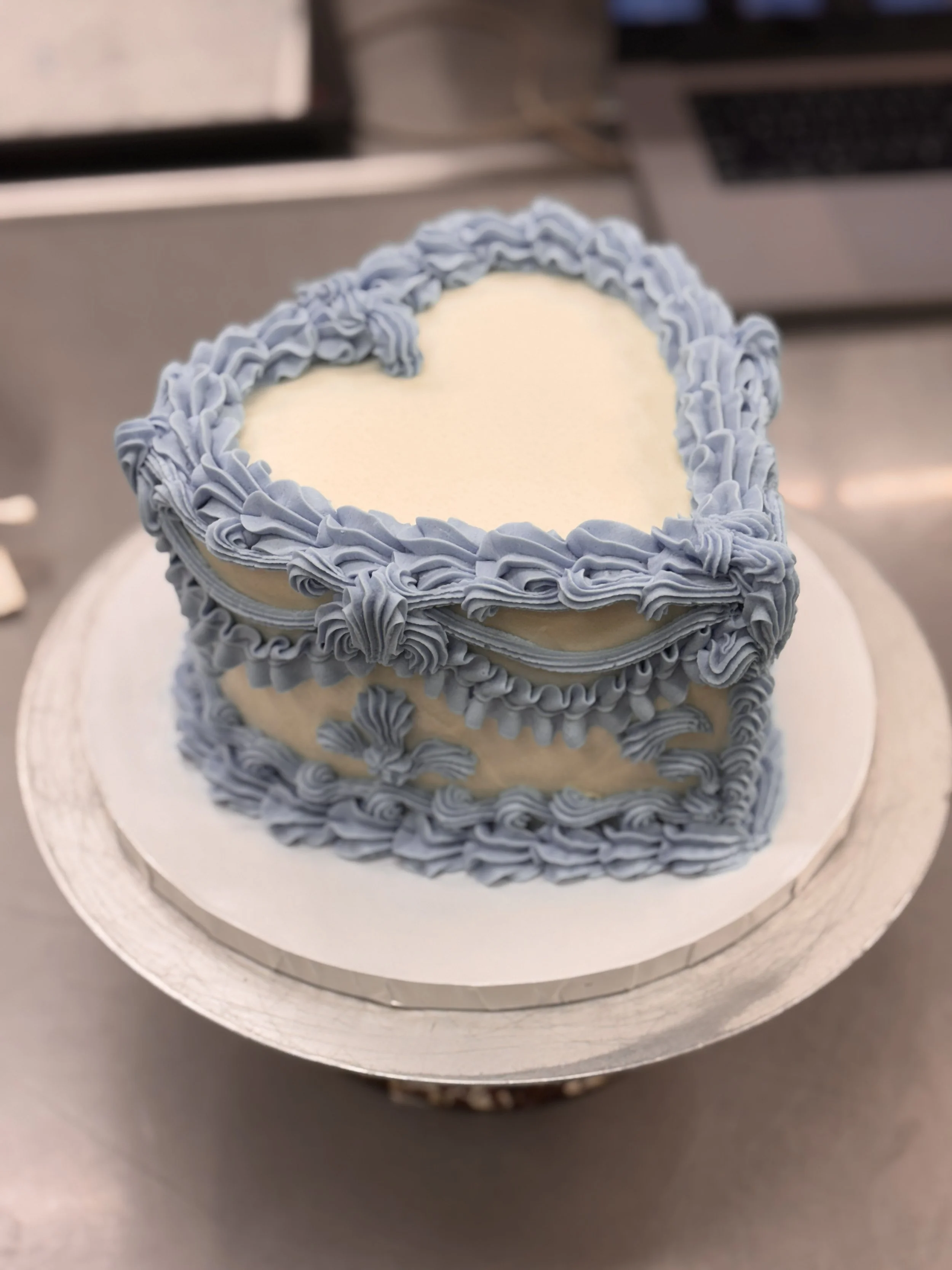 A heart-shaped cake with blue frosting decorations along the edges, sitting on a white lace cake board.