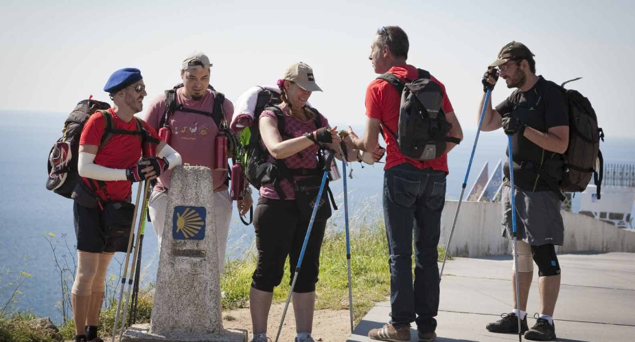 A group of five hikers standing on a trail near the ocean, exchanging something in their hands, with rugged terrain and a cloudy sky in the background.