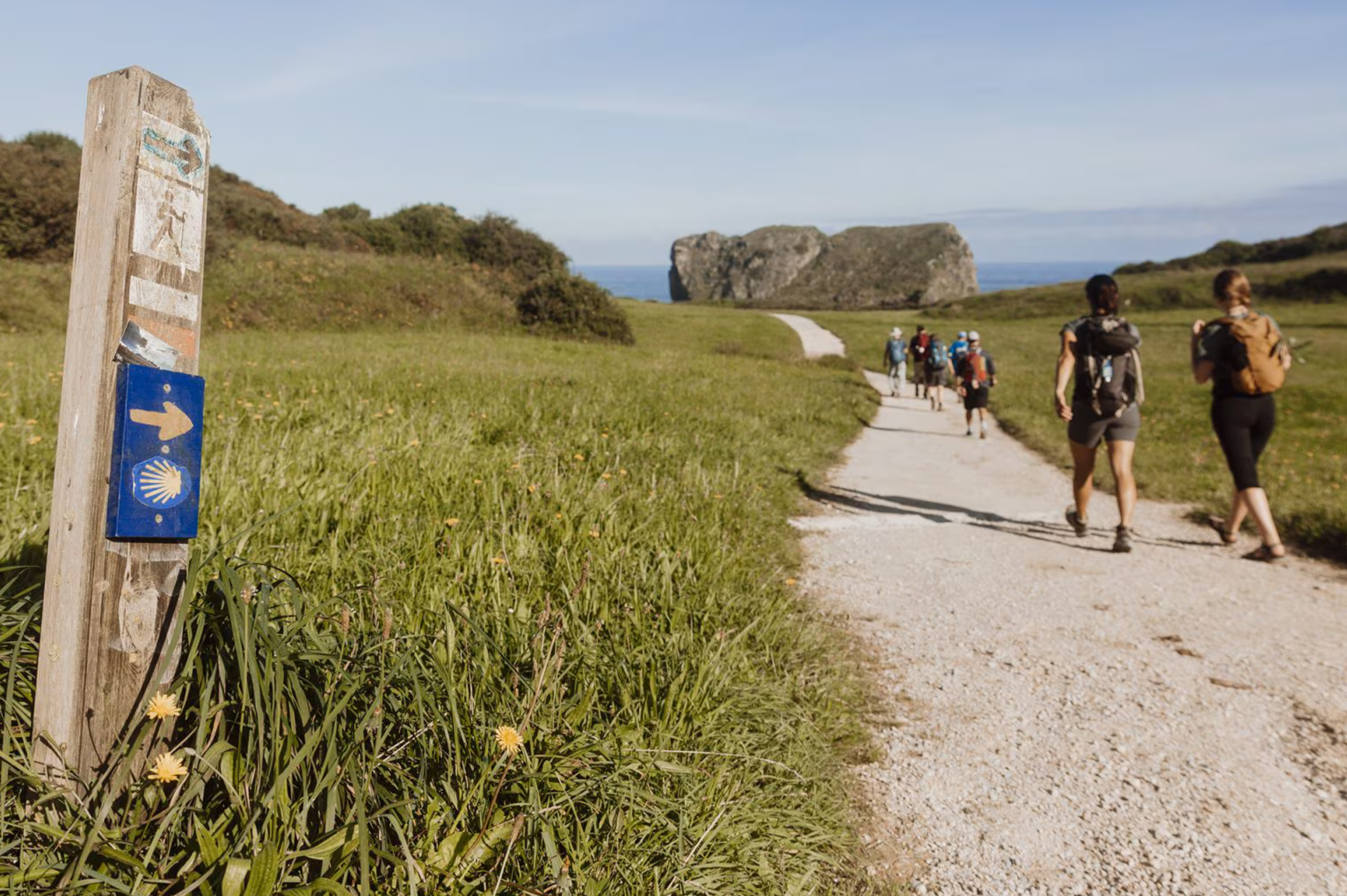 Group of hikers walking on a dirt trail in a grassy landscape with rock formations in the background. A blue trail marker with a shell symbol and an arrow is visible in the foreground.