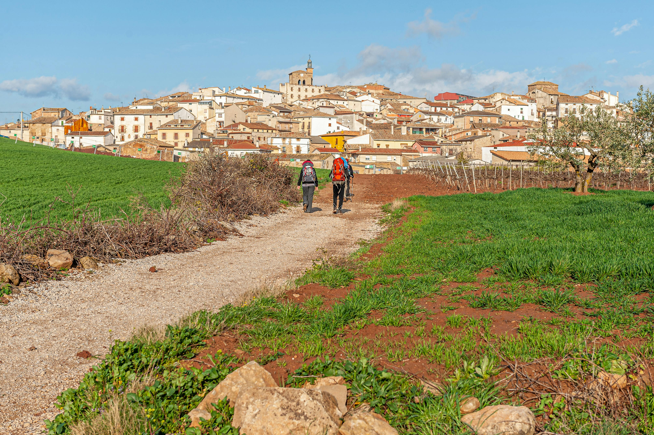 Two hikers with backpacks walking along a dirt path toward a village on a sunny day with blue sky and clouds.