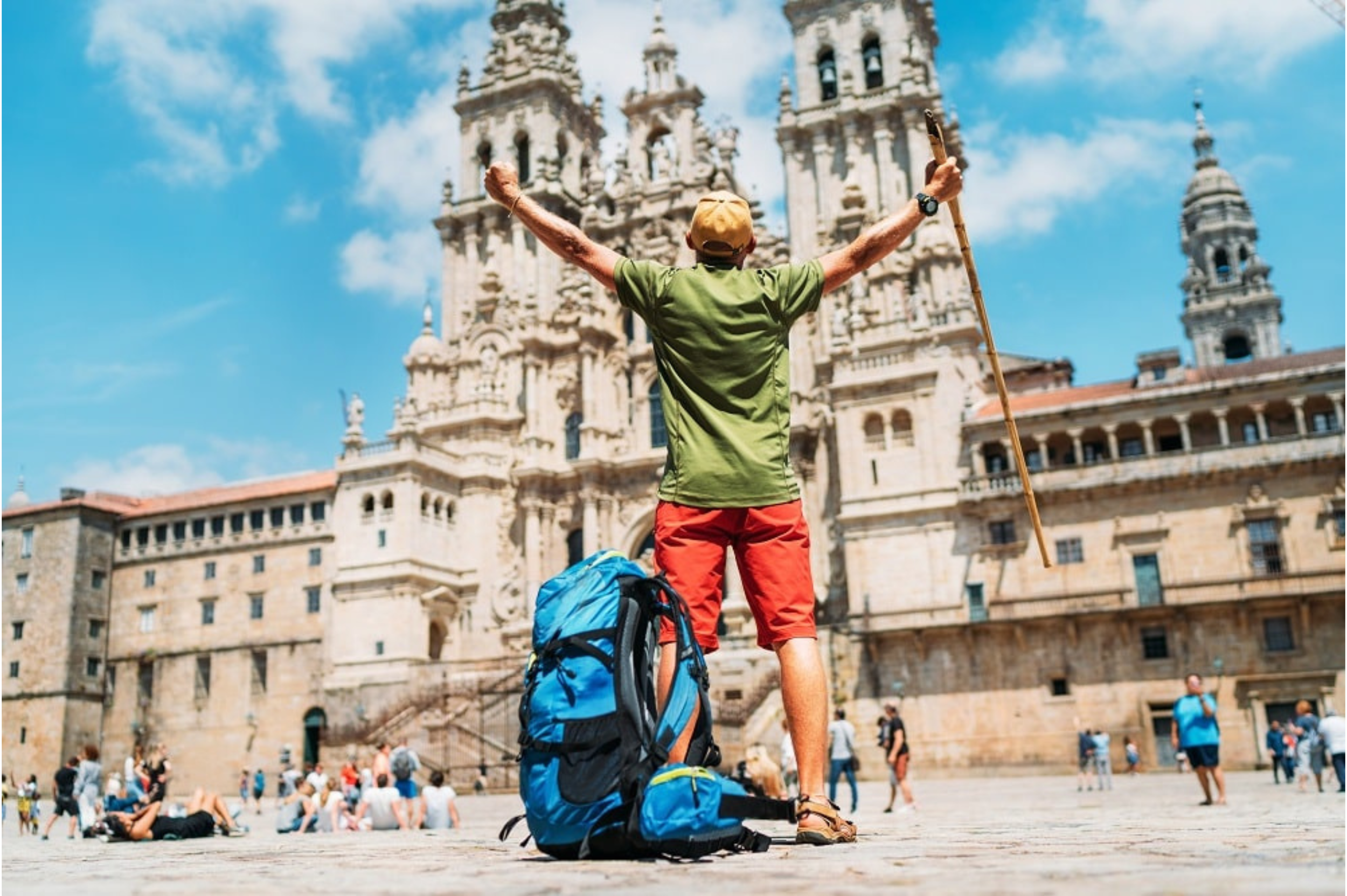 A man with a backpack standing with arms raised in front of a historic cathedral, possibly celebrating or posing for a photo, in a busy plaza with other tourists.
