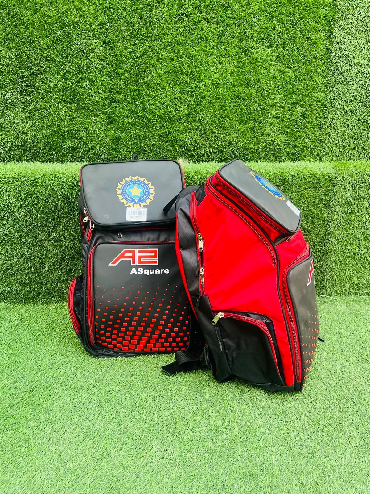 Two cricket bags, one black and red with the label 'A2 ASquare', and one black with a blue and yellow logo, resting on vibrant green artificial grass with a trimmed hedge background.