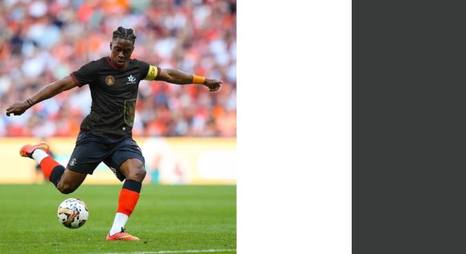 A soccer player kicking a ball on the field during a match, wearing a black jersey, red socks, and an armband, with a crowd in the background.