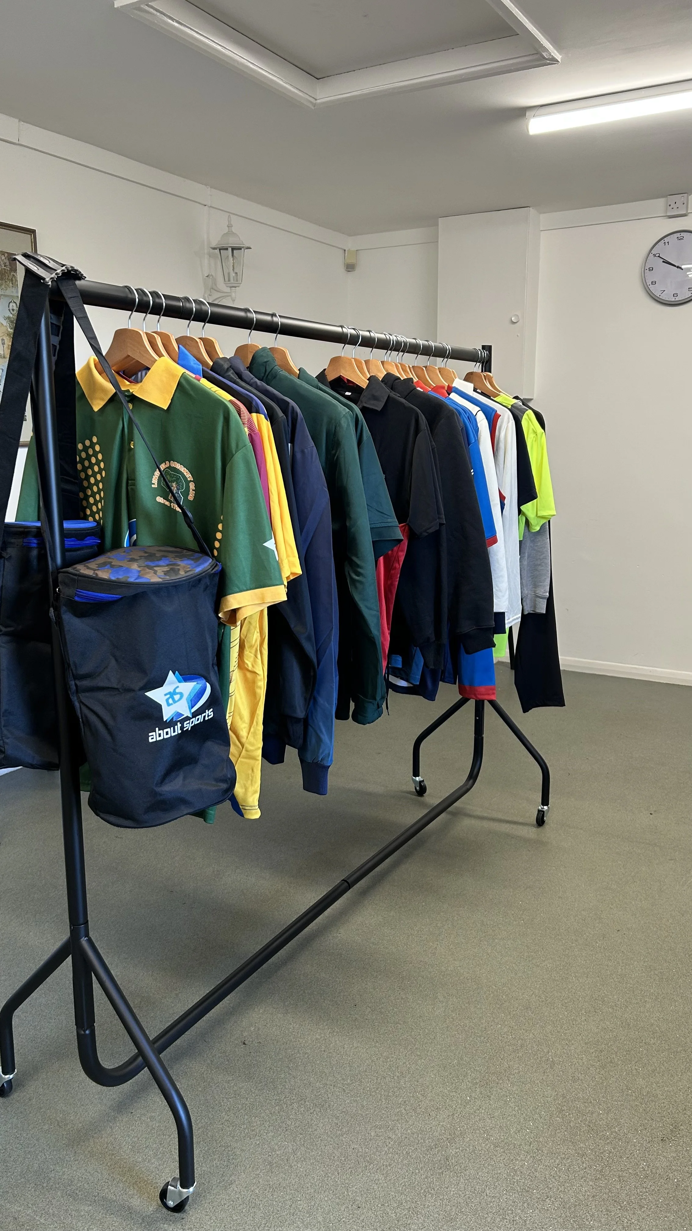 A clothing rack holding various sports jerseys and jackets in different colors, along with a black sports bag hanging from the left side of the rack. The scene is indoors with a simple, undecorated room, gray carpet, and a clock on the wall showing approximately 10:12.