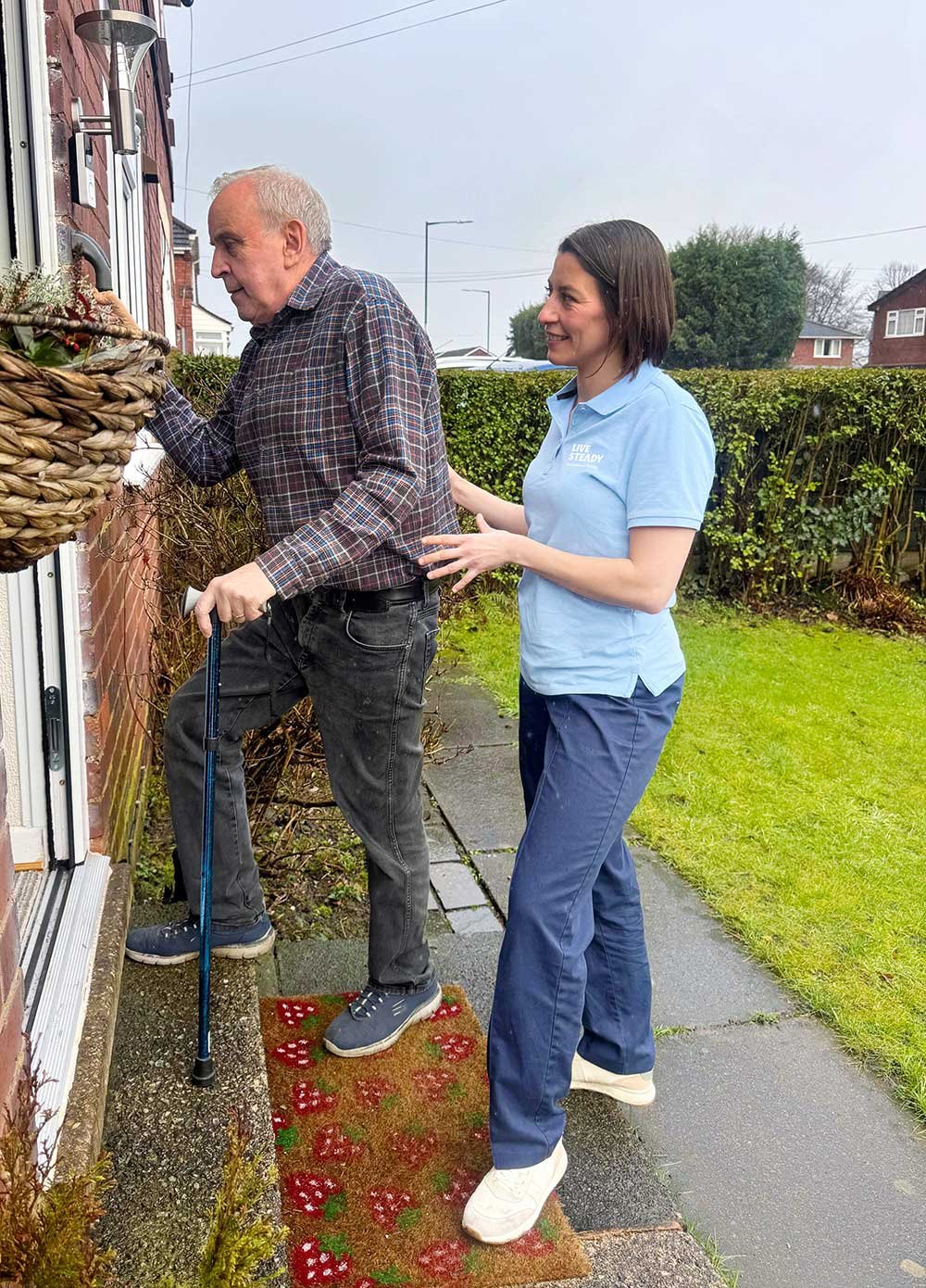 A woman with a blue shirt helping a man with a cane step onto a house's porch. The man is wearing a checkered shirt and gray pants, and the woman is standing on a decorative red and green doormat.