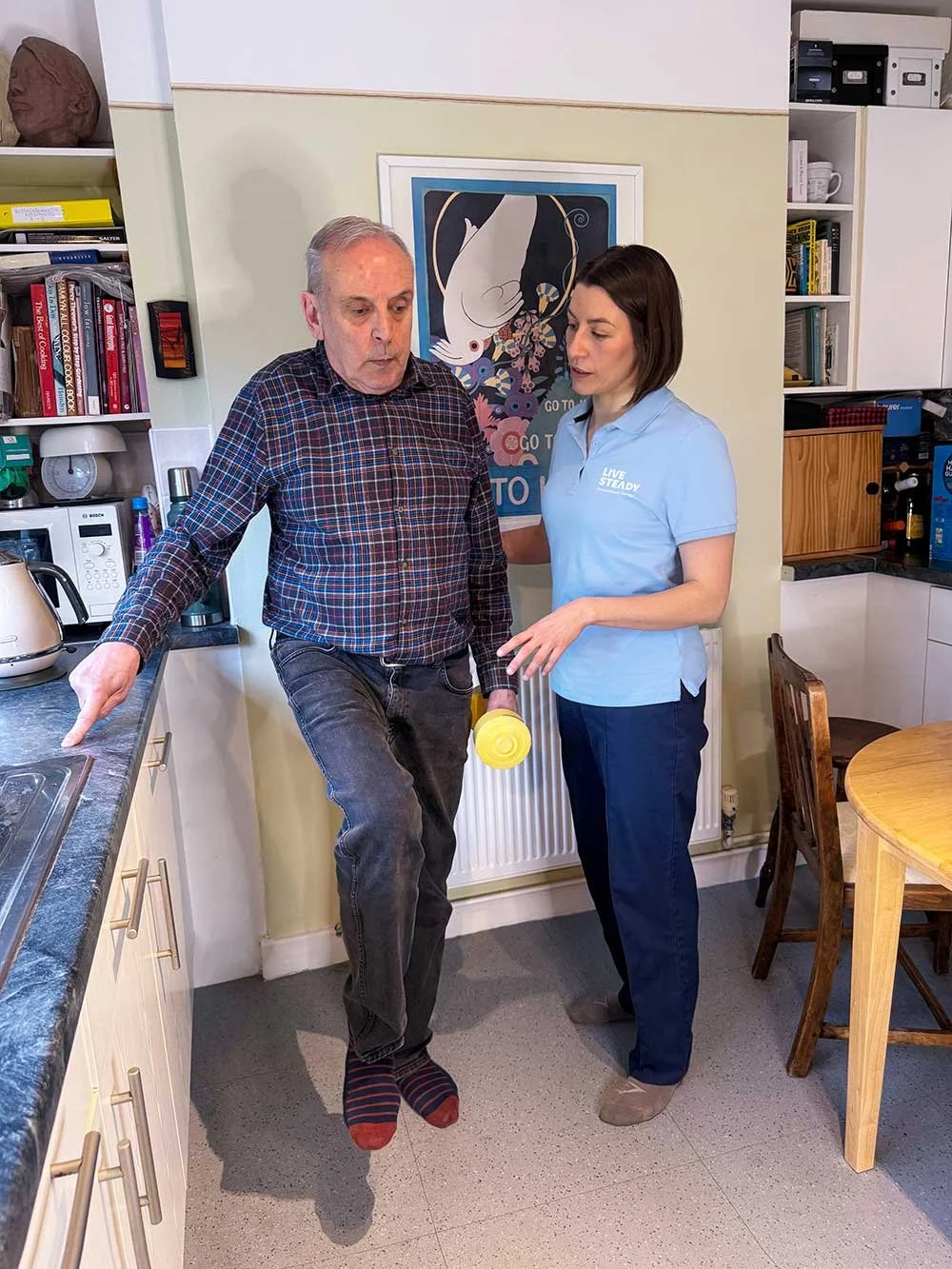A man and woman in a kitchen having a conversation. The man is wearing a checked shirt and dark jeans, standing on his tiptoes with one foot raised, and holding a yellow container. The woman is dressed in a blue polo shirt with the words 'LIVE STEADY' and blue pants, standing close to the man, looking at him and speaking. The kitchen has white cabinets, a coffee machine, a microwave, and shelves filled with books and boxes.
