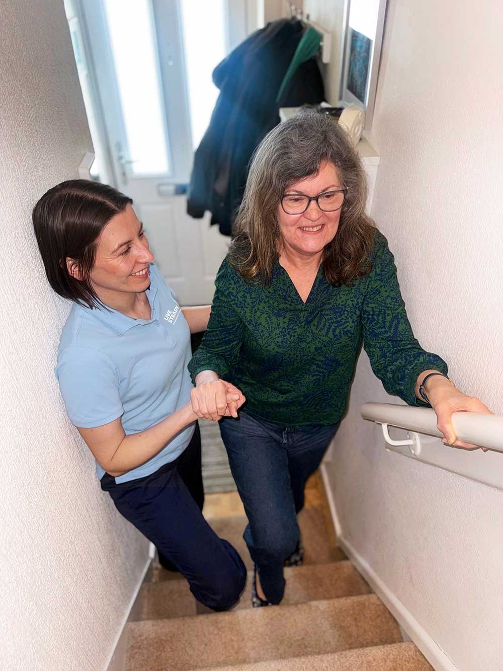 A woman assists another woman up a carpeted staircase while holding her hand. The woman at the top is wearing glasses and a green patterned shirt, and the woman below is wearing a light blue shirt. They are smiling and appear to be in a home or residential setting.