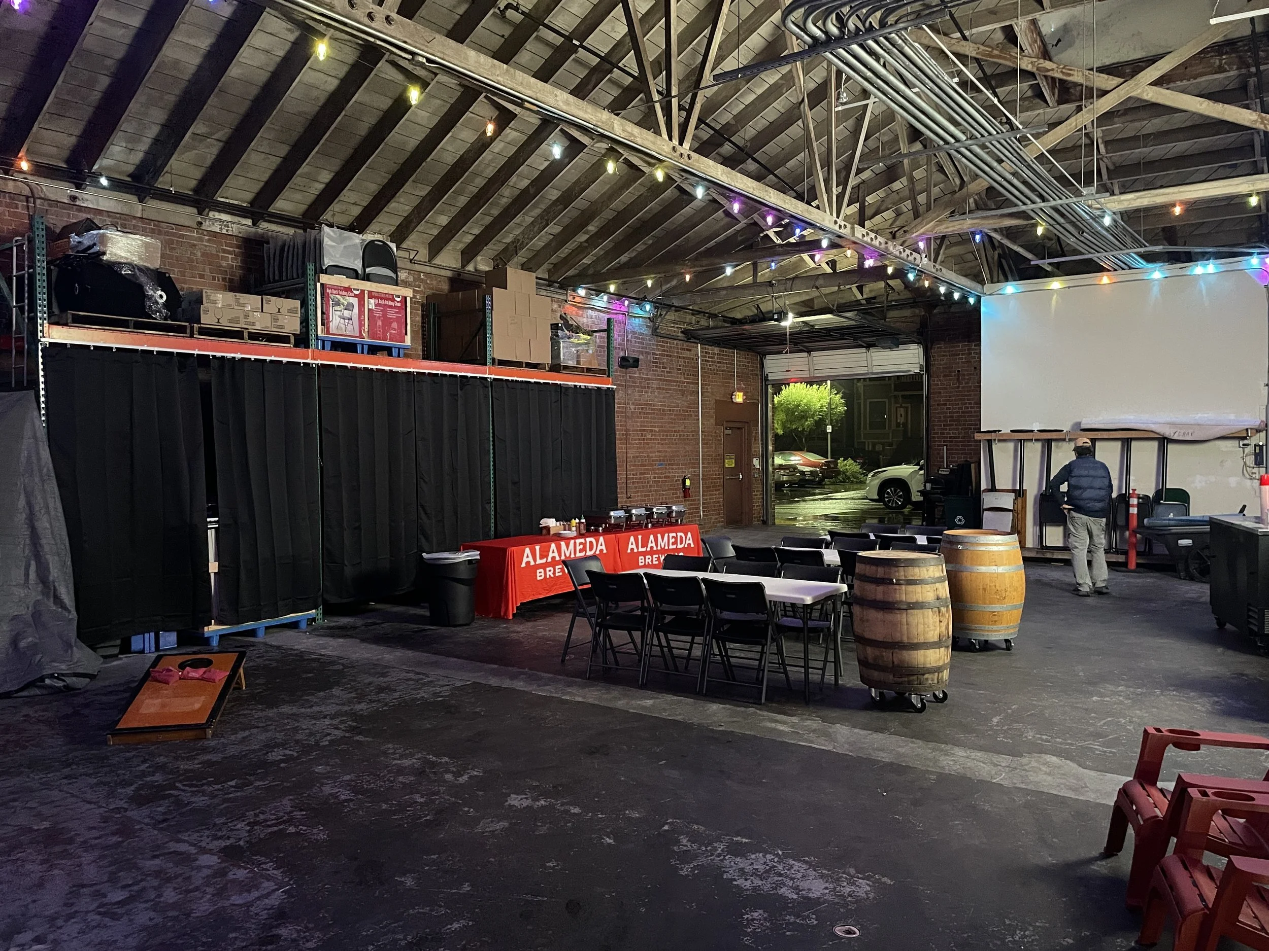 Empty indoor event space with tables, wine barrels, and festival lighting; a man stands near the door outside on a rainy night.
