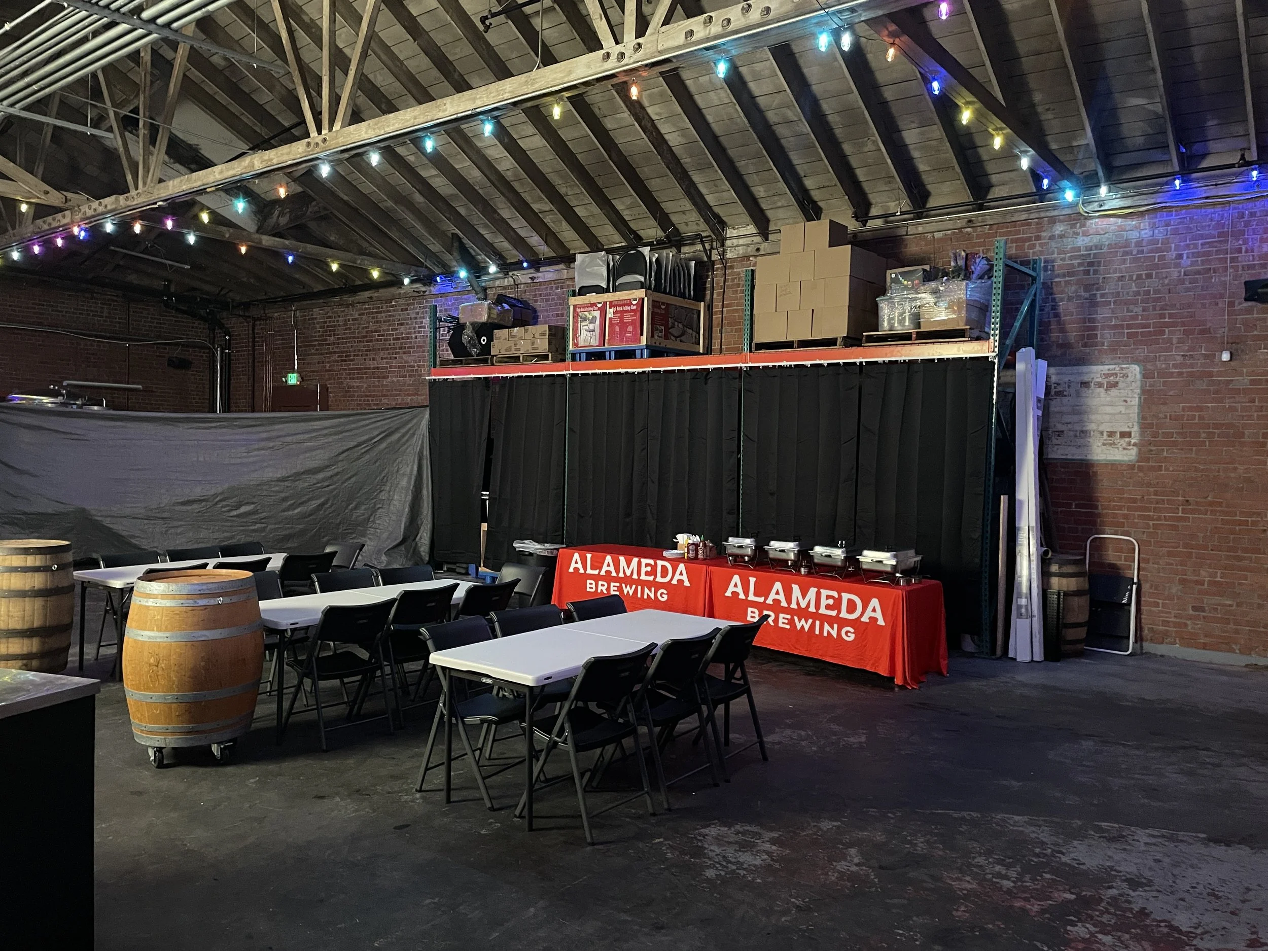 Indoor event space with tables and chairs, decorated with colorful string lights, and a table with 'Alameda Brewing' printed on red covers, set up for serving food or drinks.