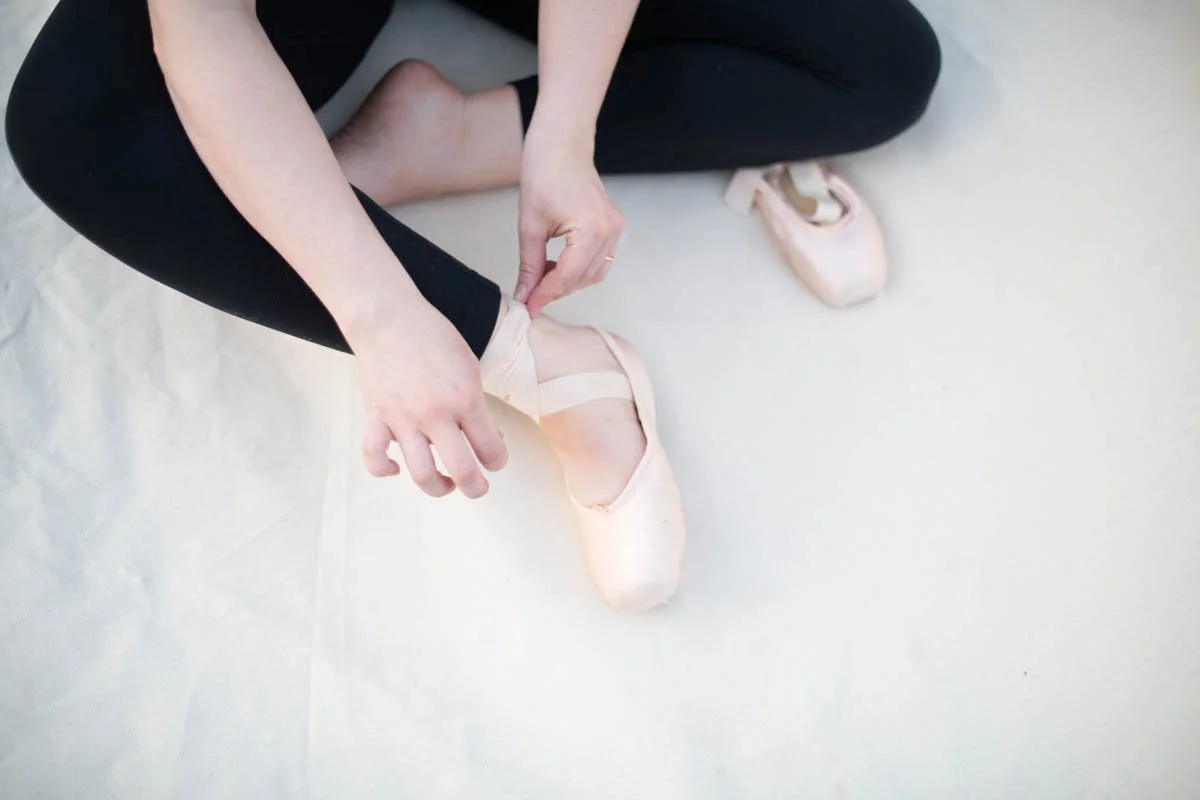 A person sitting on the floor, tying the ribbons of ballet pointe shoes, wearing black leggings.