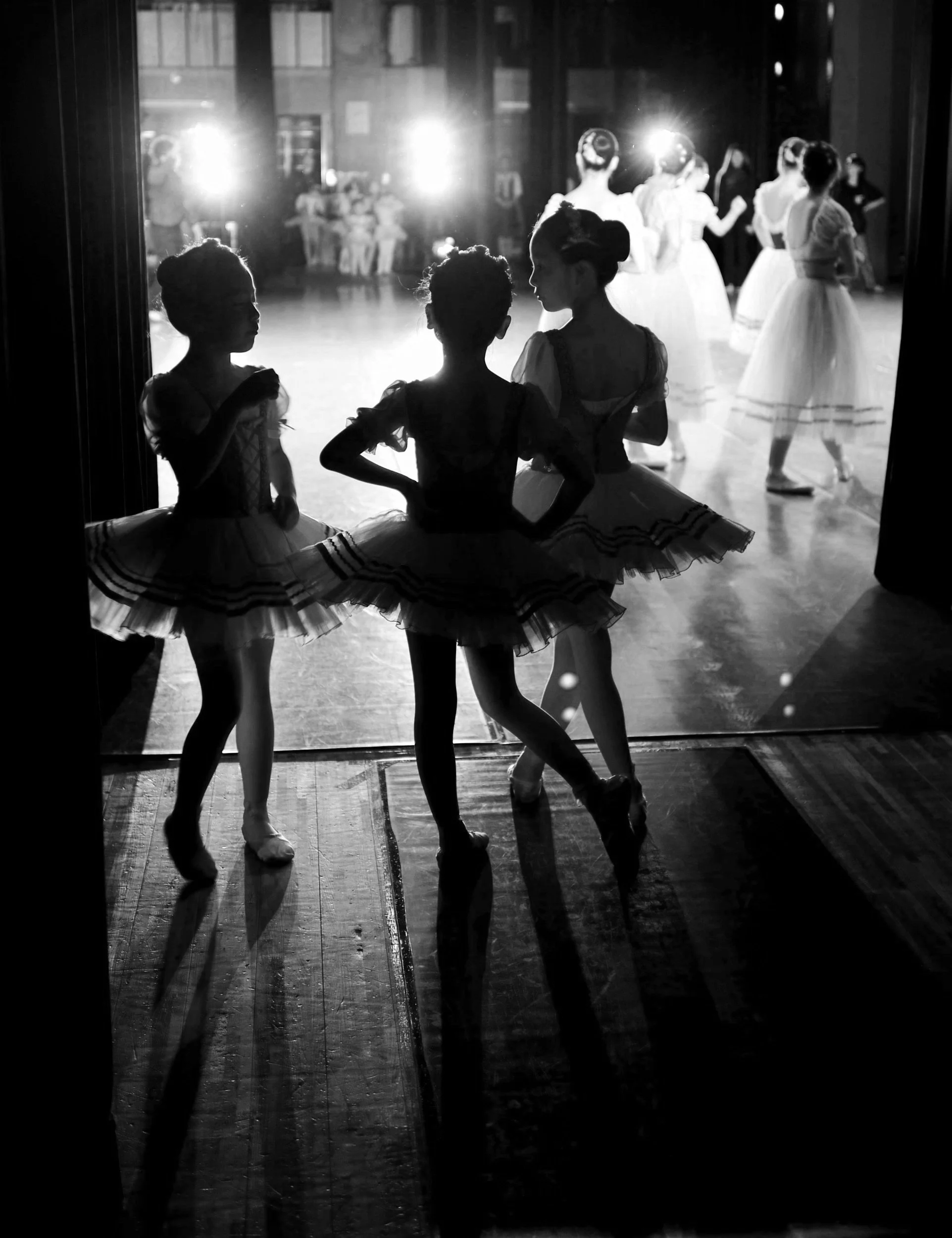 Three young ballerinas in ballet costumes standing in a doorway, silhouetted against a brightly lit dance studio with other dancers practicing in the background.