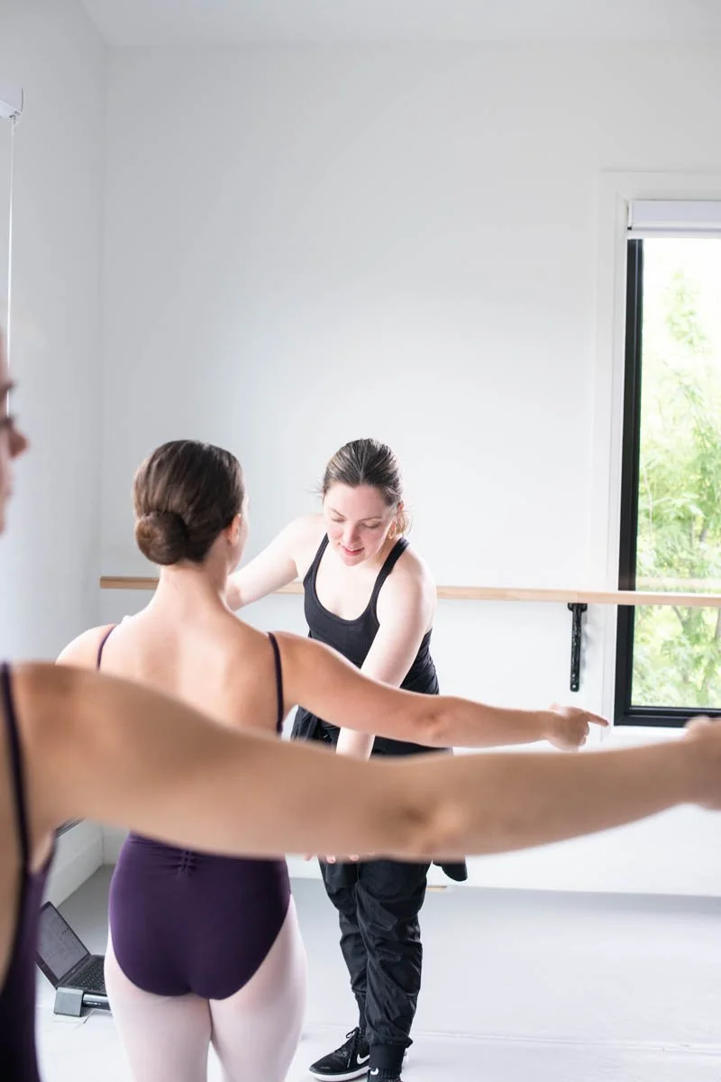 Ballet instructor assisting two students during a class in a bright studio with a window and ballet bar.
