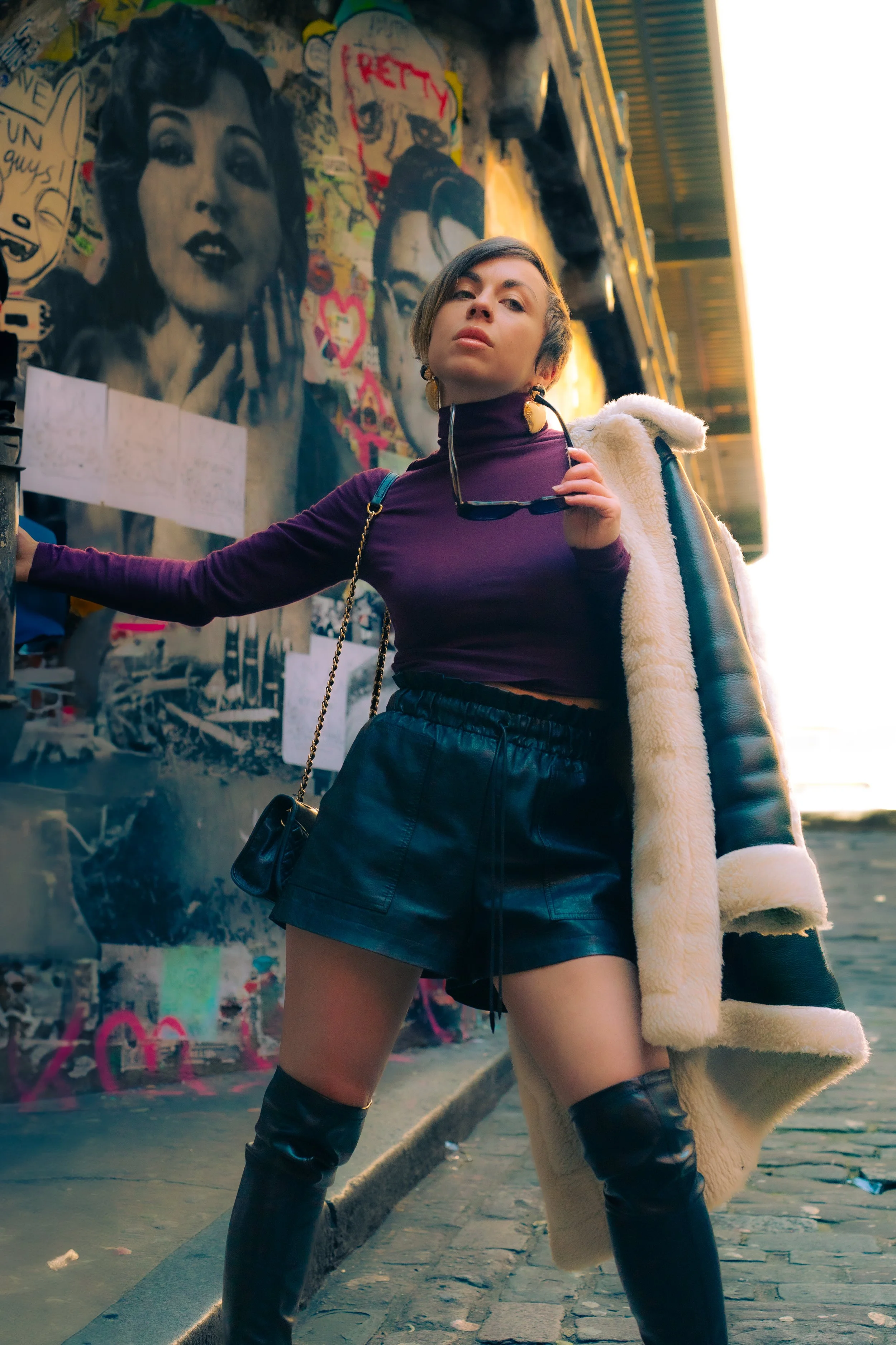 A person in fashionable attire posing on a street with urban graffiti art in the background.