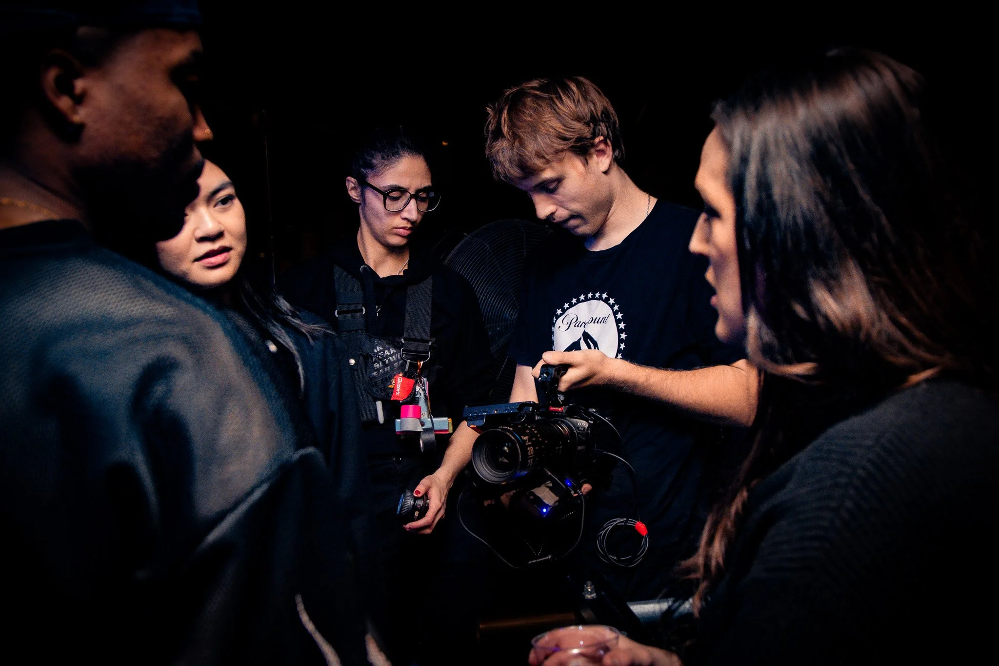 Group of people in a dimly lit setting focused on a person operating a film camera, wearing casual clothing.