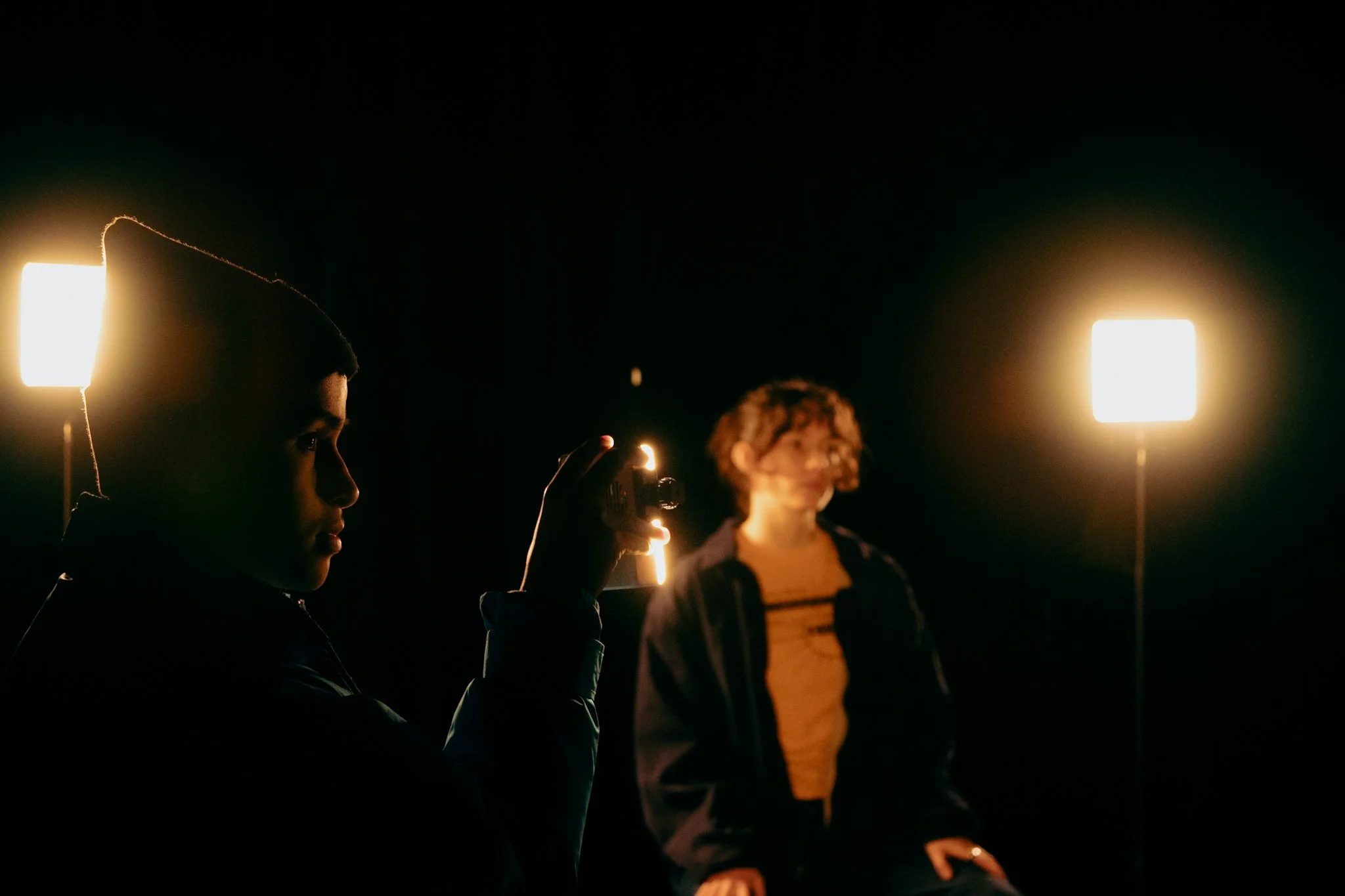 Two people in a dark room with bright square lights; one is holding a camera, and a woman with curly hair and glasses sits in front of the lights.