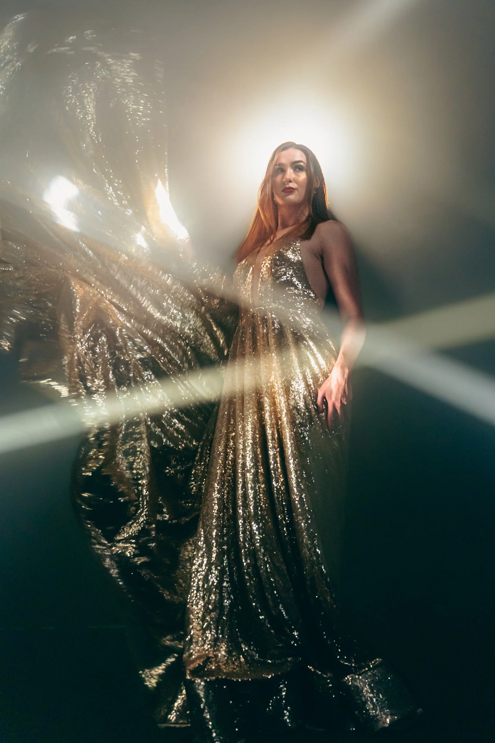 A woman in a shimmering, gold, sequined evening gown standing amidst fog or smoke with bright spotlights creating a halo effect behind her, and light flares across the image.