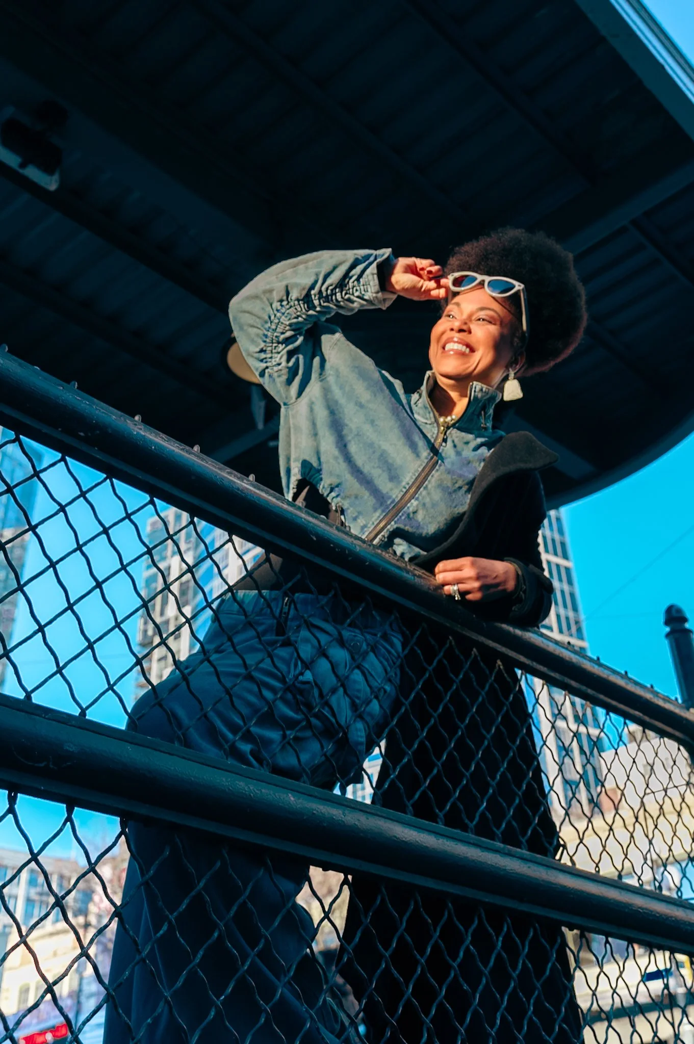 A smiling woman with an afro hairstyle, sunglasses, and earrings standing on a city balcony with a metal railing, looking into the distance under a dark overpass with tall buildings in the background.
