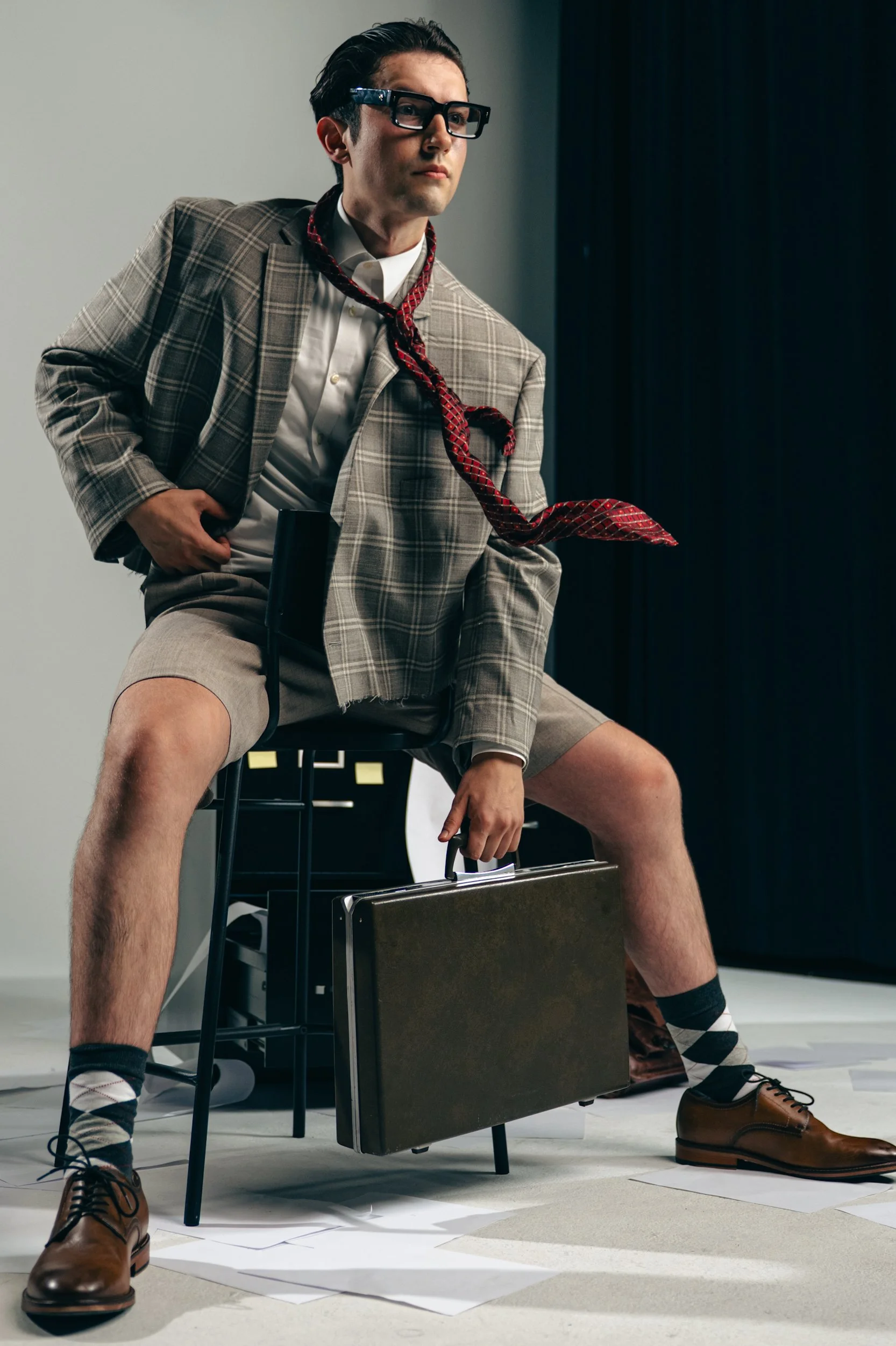 Young man dressed in a blazer, shorts, and dress shoes, sitting on a chair with a briefcase, surrounded by scattered papers, and wearing glasses with a loosened shirt collar and a necktie.