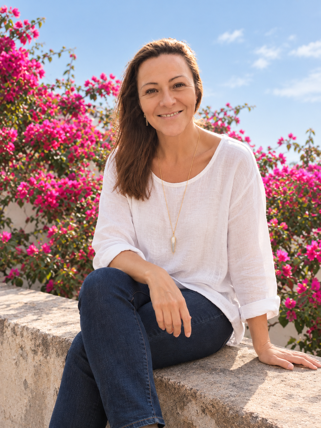Mujer sonriendo sentada en una roca con flores rosadas de fondo y cielo azul claro.