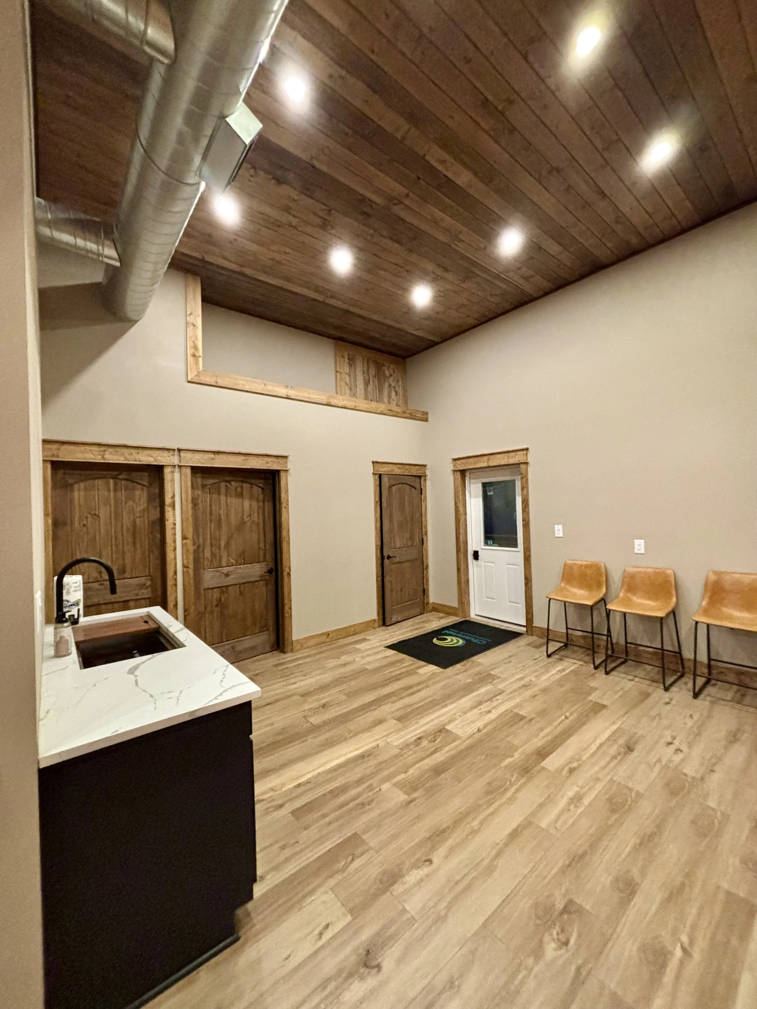 Interior room with wooden ceiling and floors, three chairs along the wall, a small kitchenette with a sink, and a door with a window next to a black welcome mat.