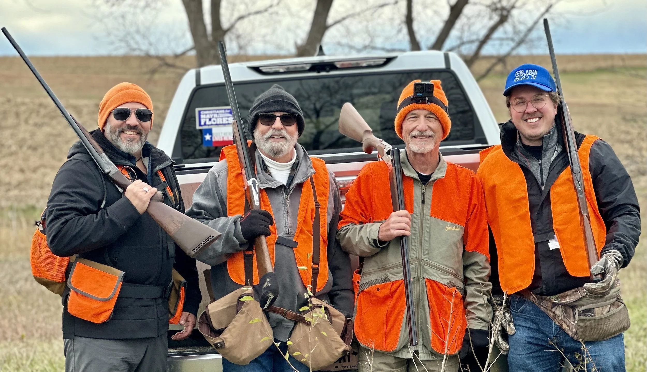 Four men outdoors standing in front of a pickup truck with a Texas flag sticker, dressed in outdoor hunting gear, holding rifles, and wearing orange safety vests and hats, smiling.