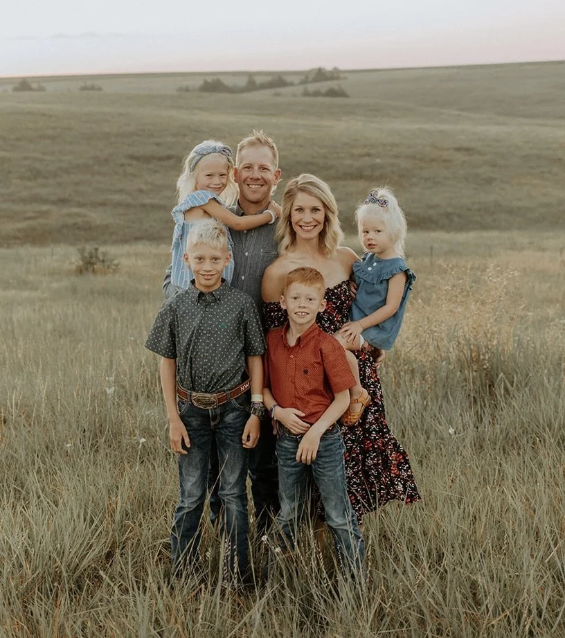 A smiling family of six outdoors in a grassy field with rolling hills in the background.