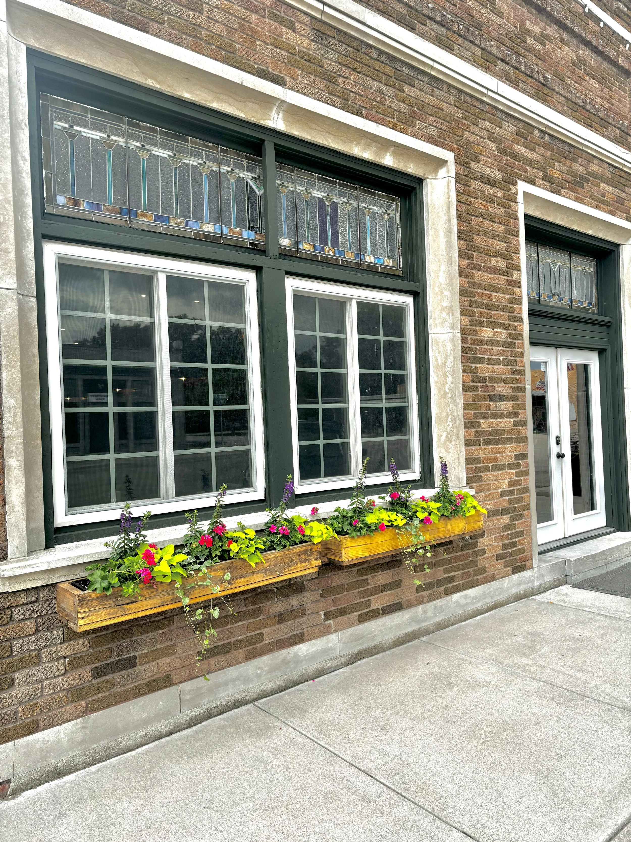 Brick building with large window with stained glass upper section, flower boxes with colorful flowers, and a doorway with glass panels.