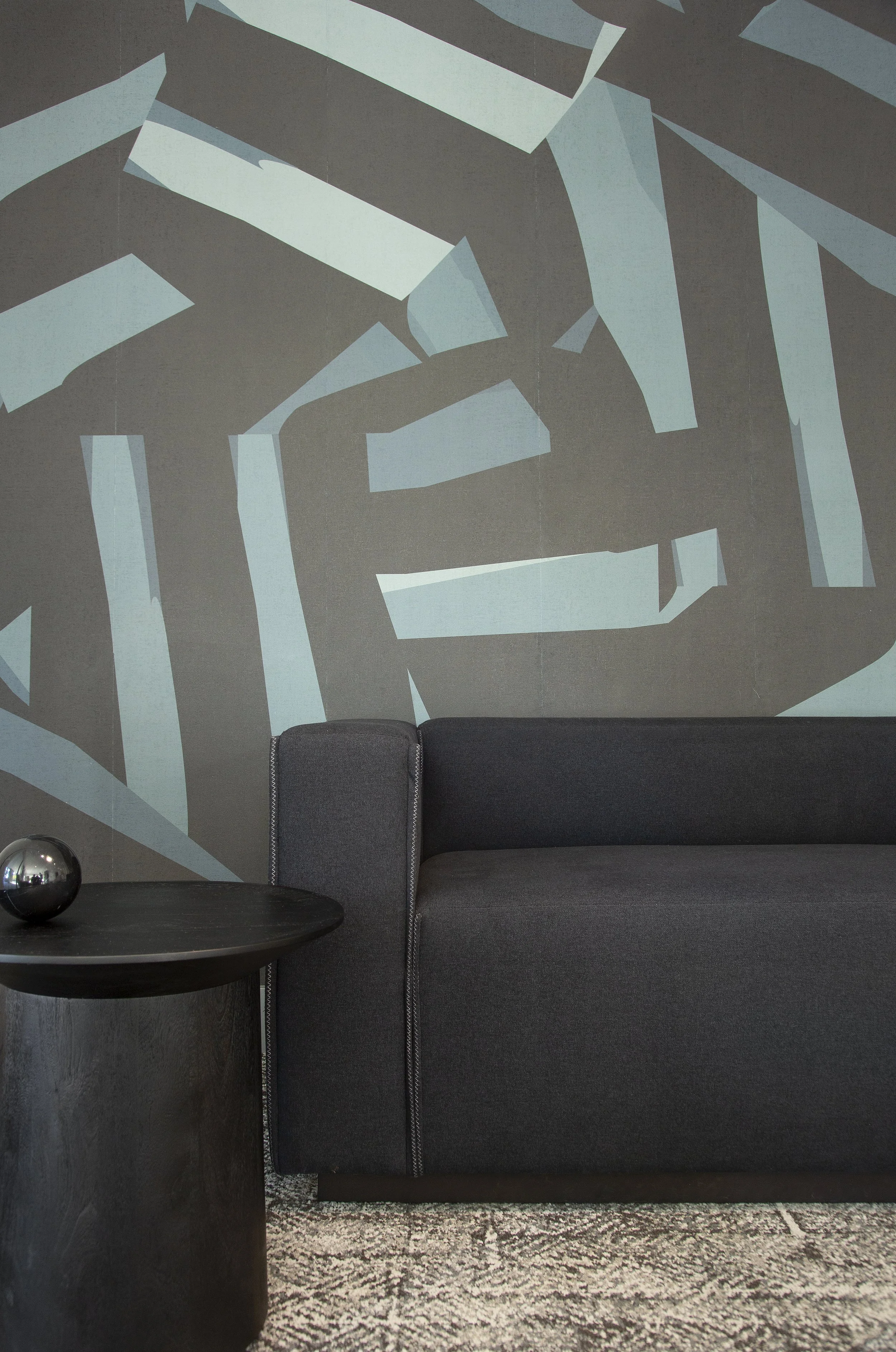 A black sofa with a dark gray backrest and a round black side table with a reflective metallic ball on top, against a wall with abstract geometric wall art in shades of blue and gray.
