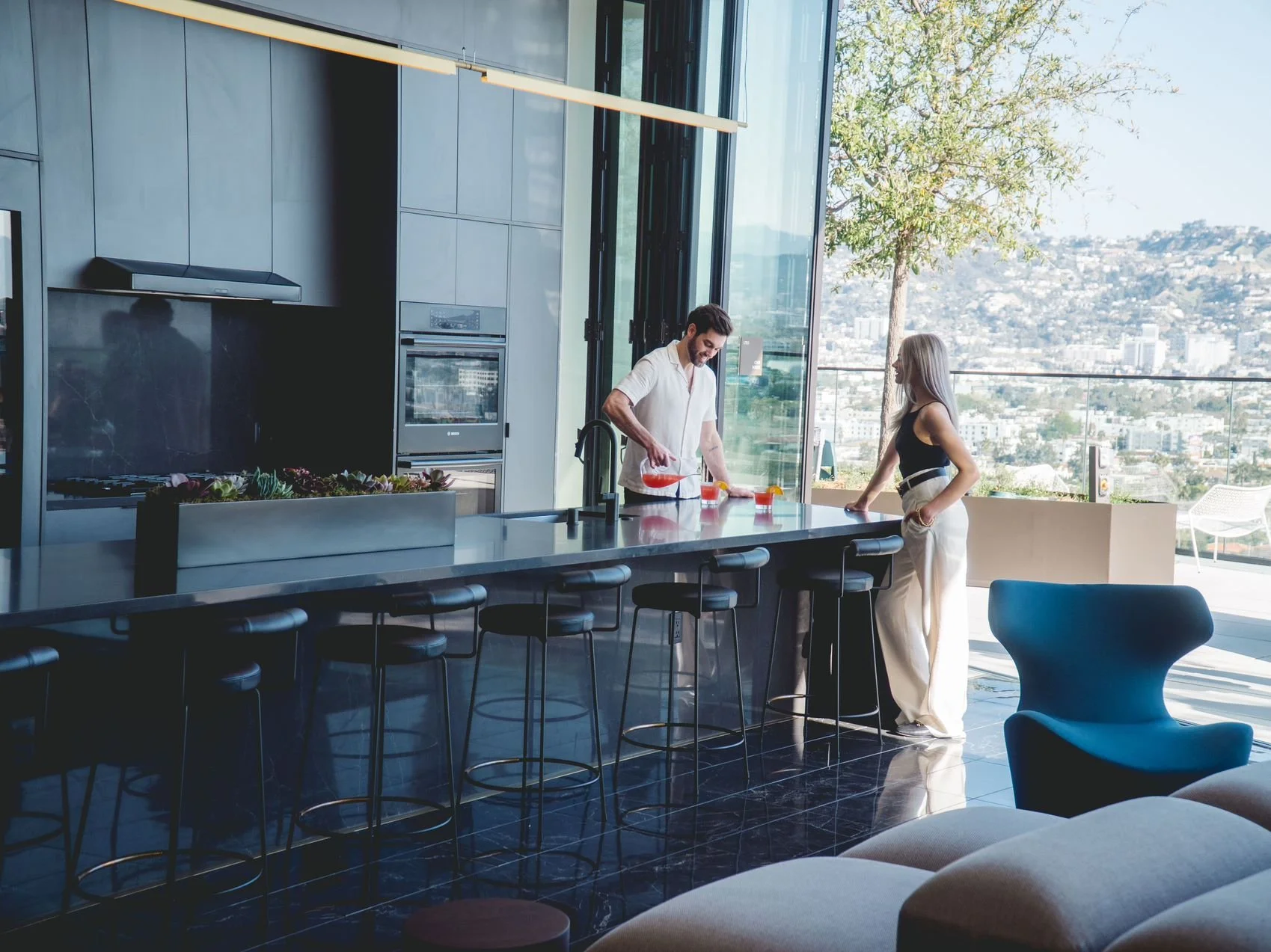 A man and woman in a modern kitchen with large windows and a city view. The man is pouring drinks at the kitchen island, and the woman is standing nearby. The kitchen has black bar stools and contemporary appliances.