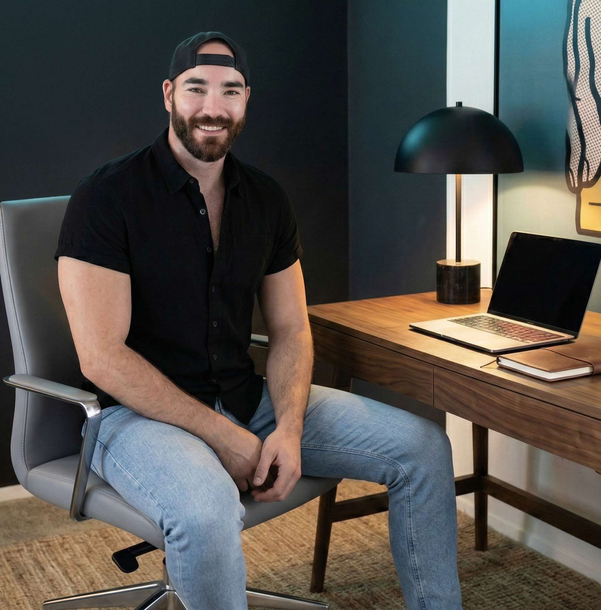 A smiling man with a beard, wearing a backward baseball cap, black short-sleeve button-up shirt, and light blue jeans, sitting on a modern office chair in a contemporary workspace. There is a wooden desk with an open laptop, a closed notebook, and a desk lamp behind him, in front of a black wall with a colorful abstract painting.