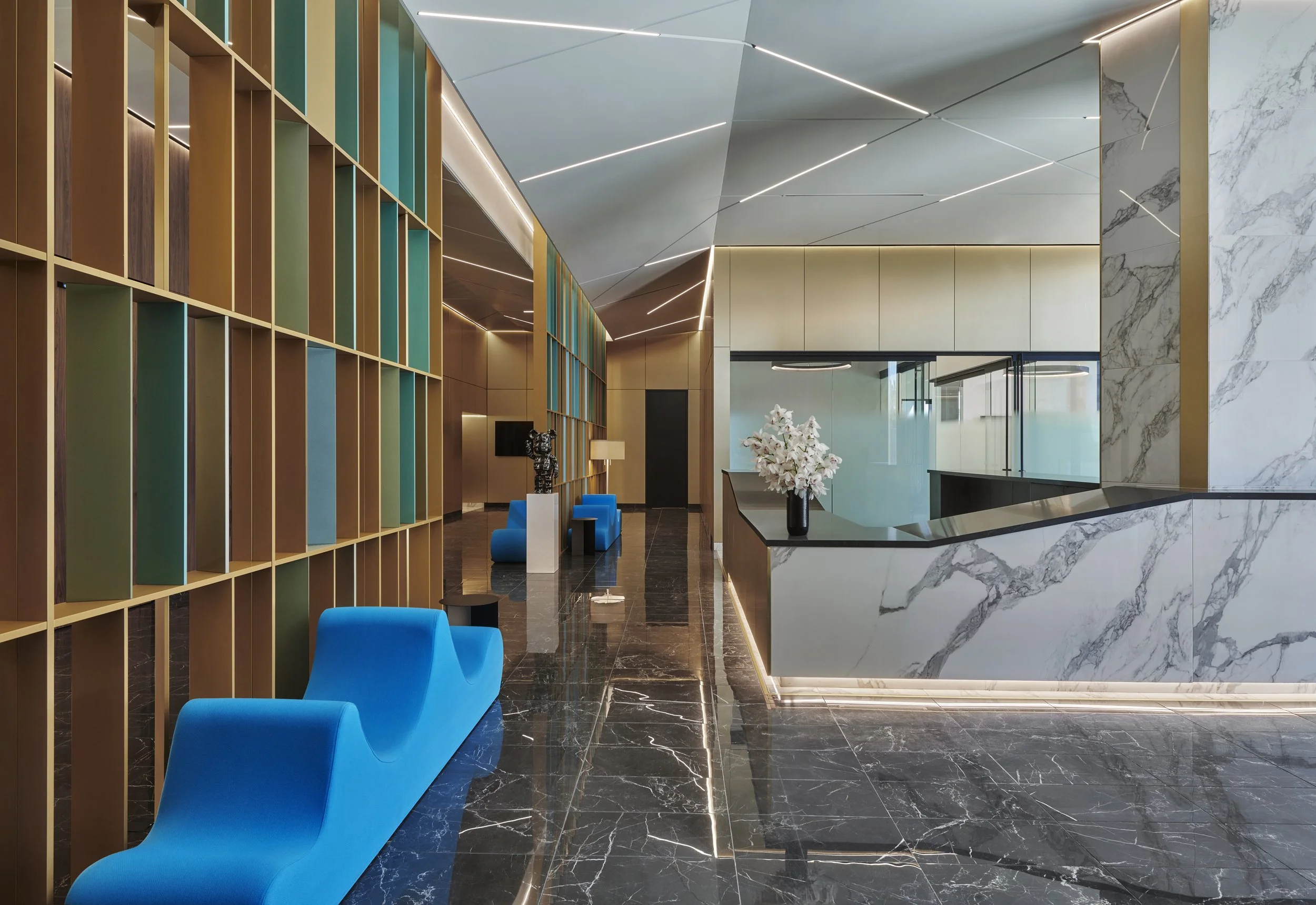Modern hotel lobby with marble floors, a colorful wall with shelves, blue chairs, sculptures, and a reception area with a flower arrangement.