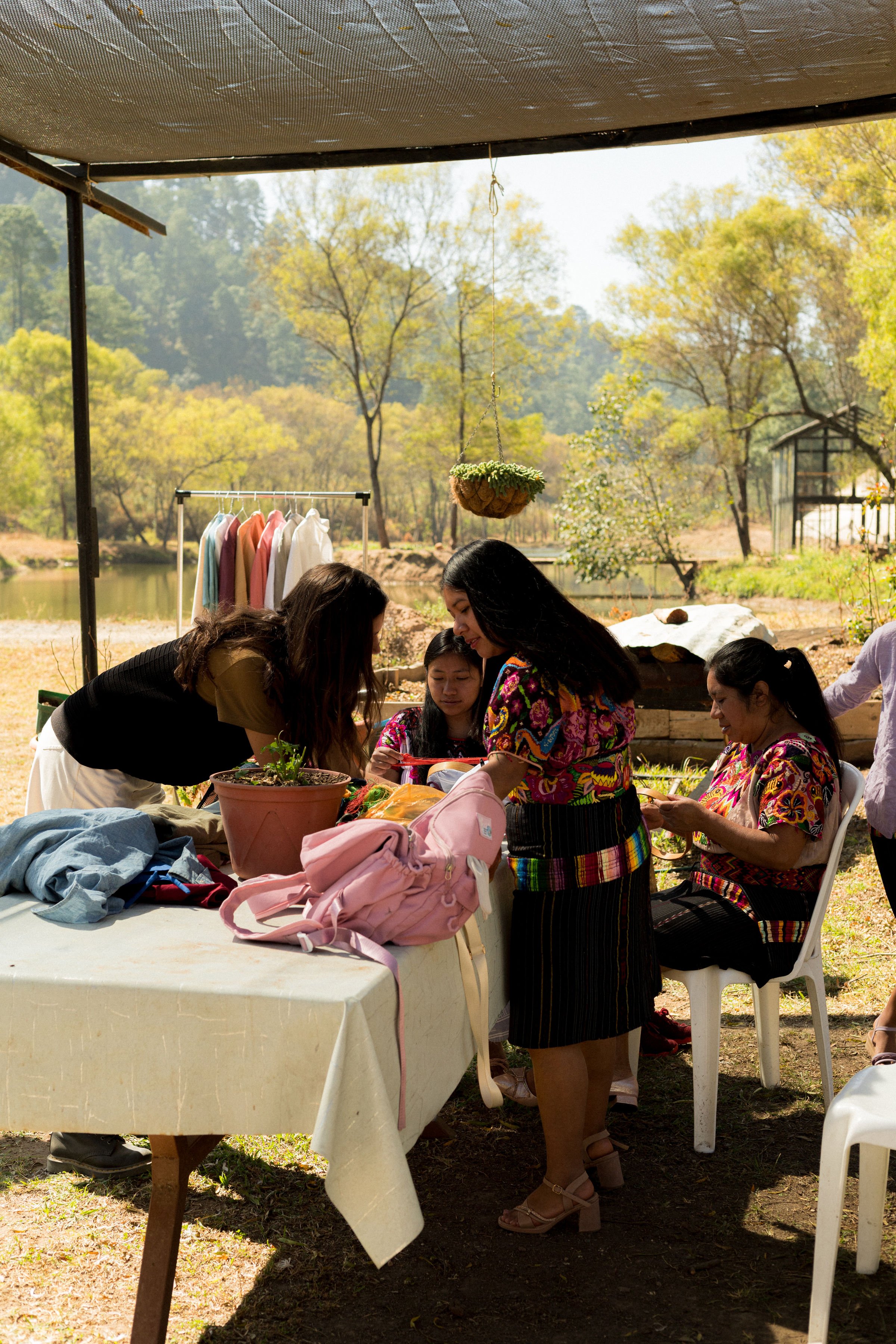 A group of women gathered around a table outdoors in a natural setting, engaged in an activity. The background features trees with changing leaves, a small lake, and a greenhouse. Clothes are hanging on a rack nearby.