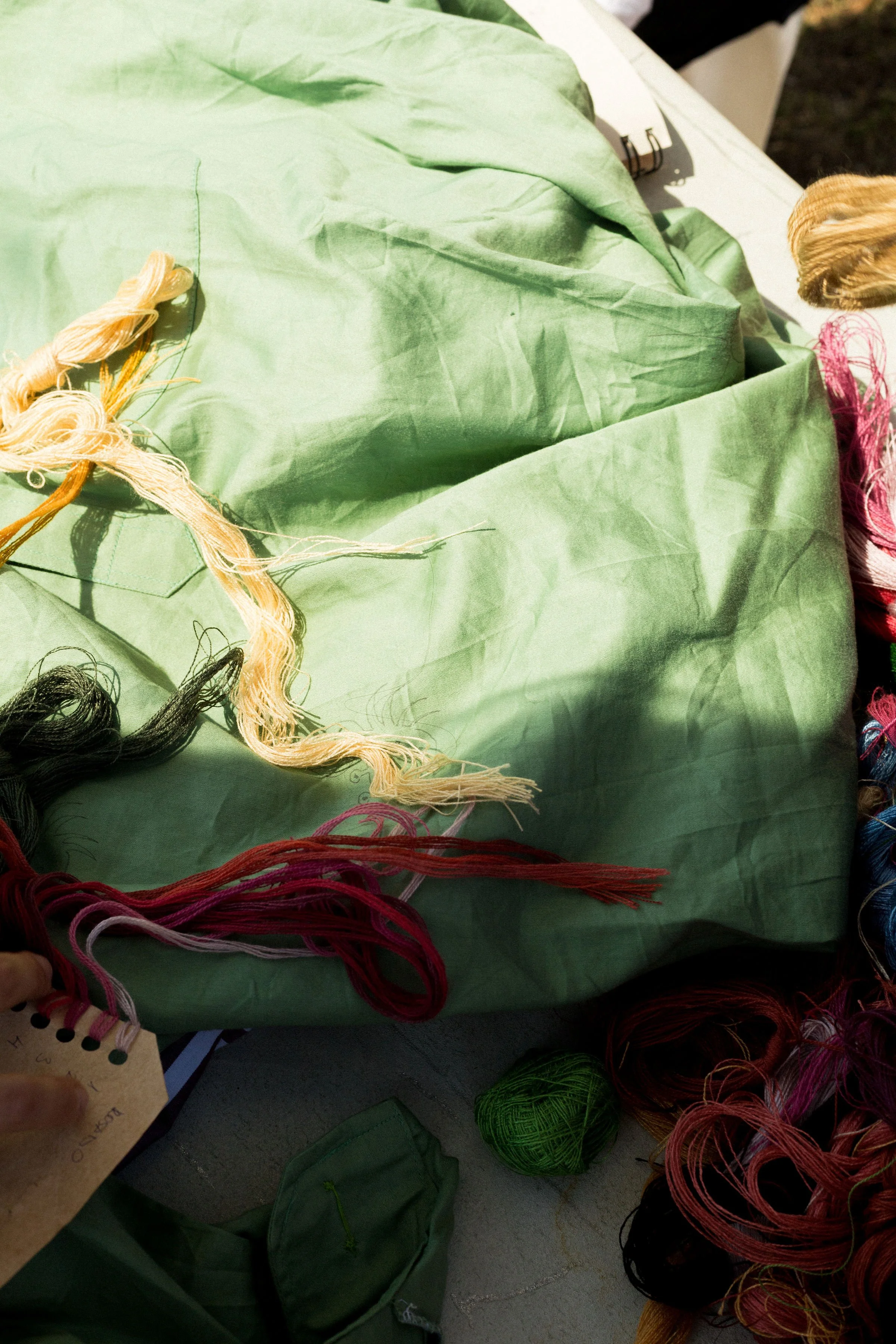 Assorted skeins of embroidery floss in various colors, including pink, black, red, green, and multicolored, on a table along with green fabric, a small pouch, and a notebook.