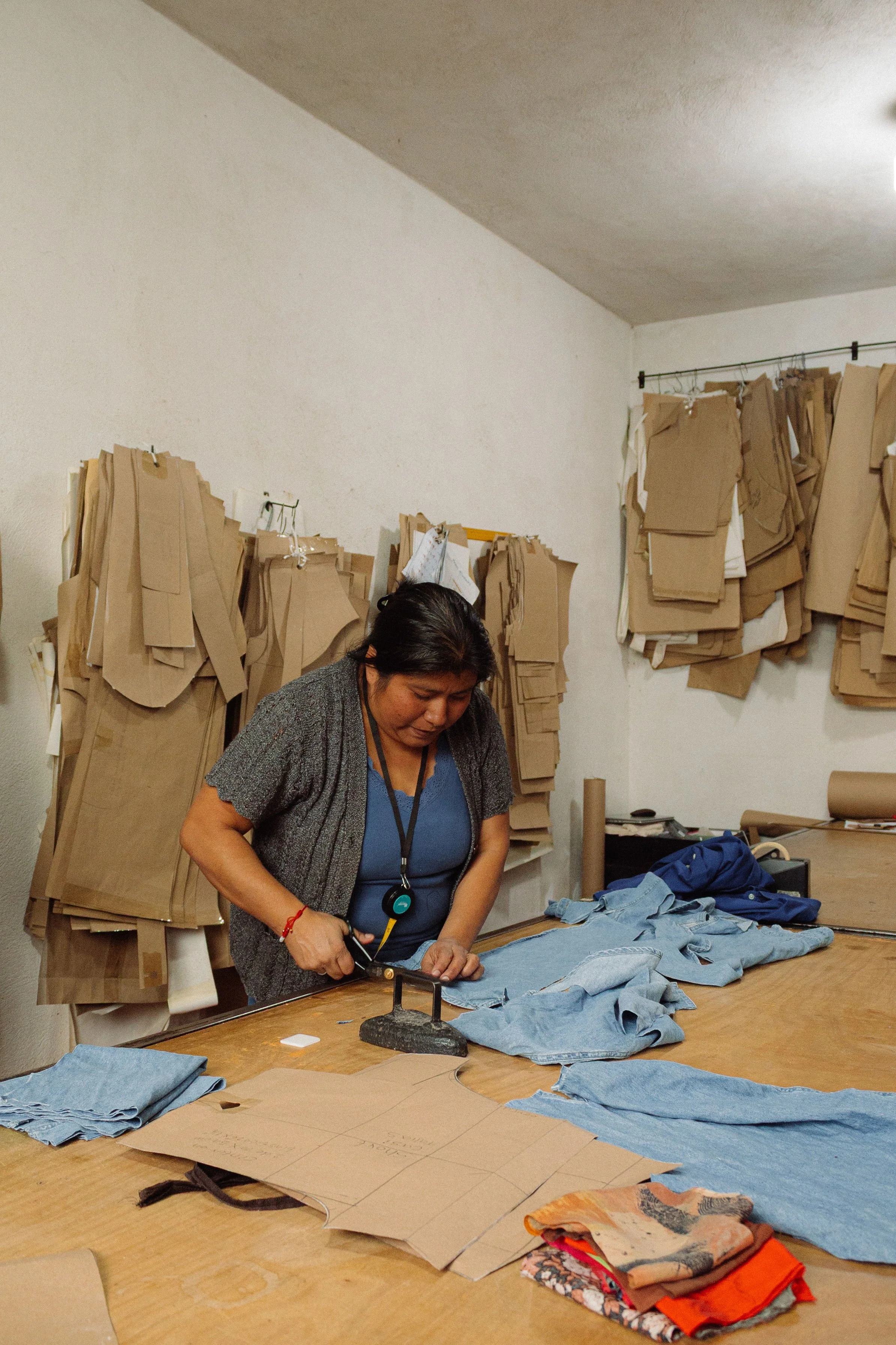 A woman working in a clothing workshop, cutting denim fabric on a table surrounded by folded jeans, patterns, and brown paper patterns on the wall behind her.
